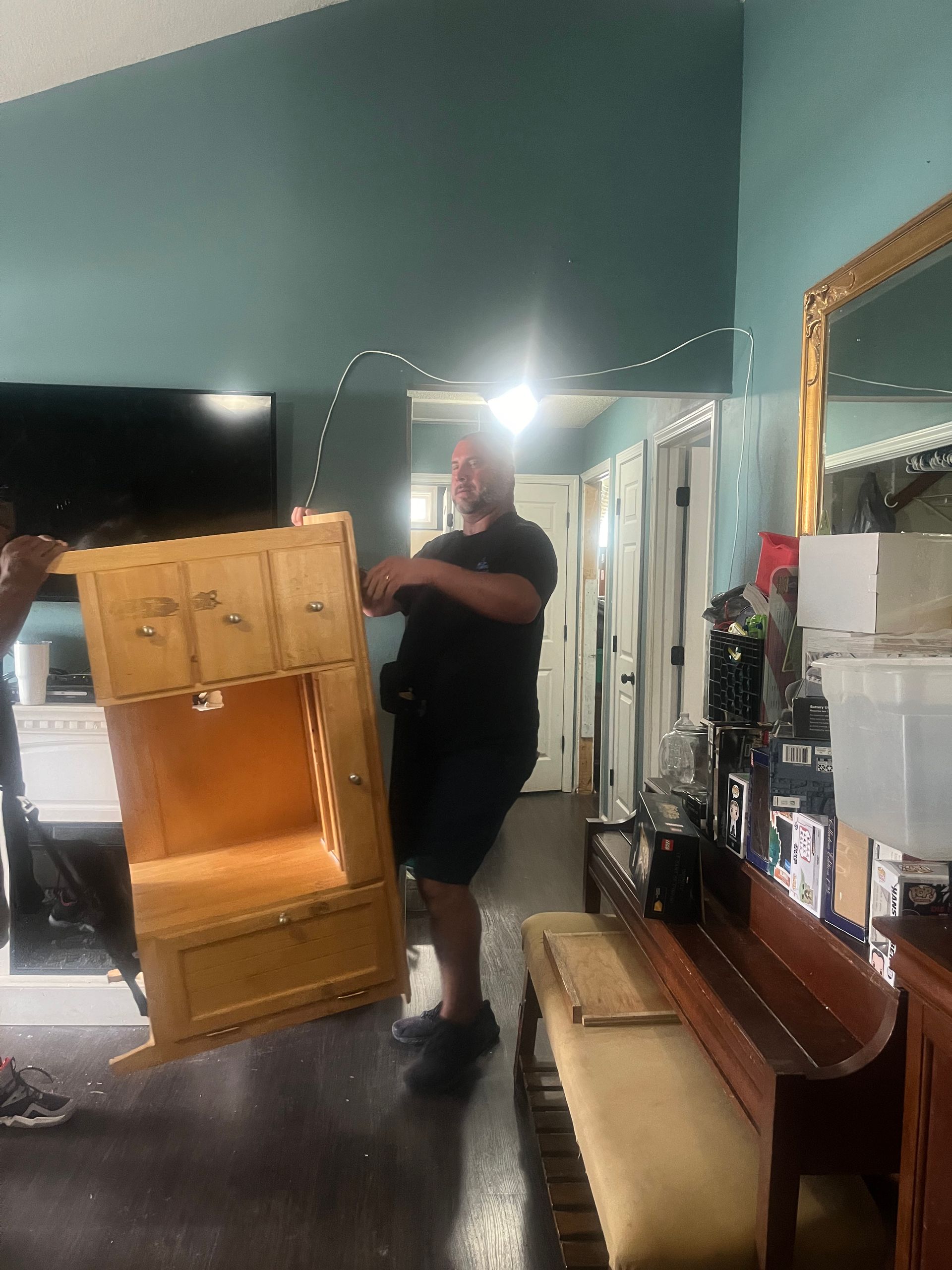 Two movers carrying a light wood cabinet. One man in black shirt and shorts, standing in a room with a piano and mirror.
