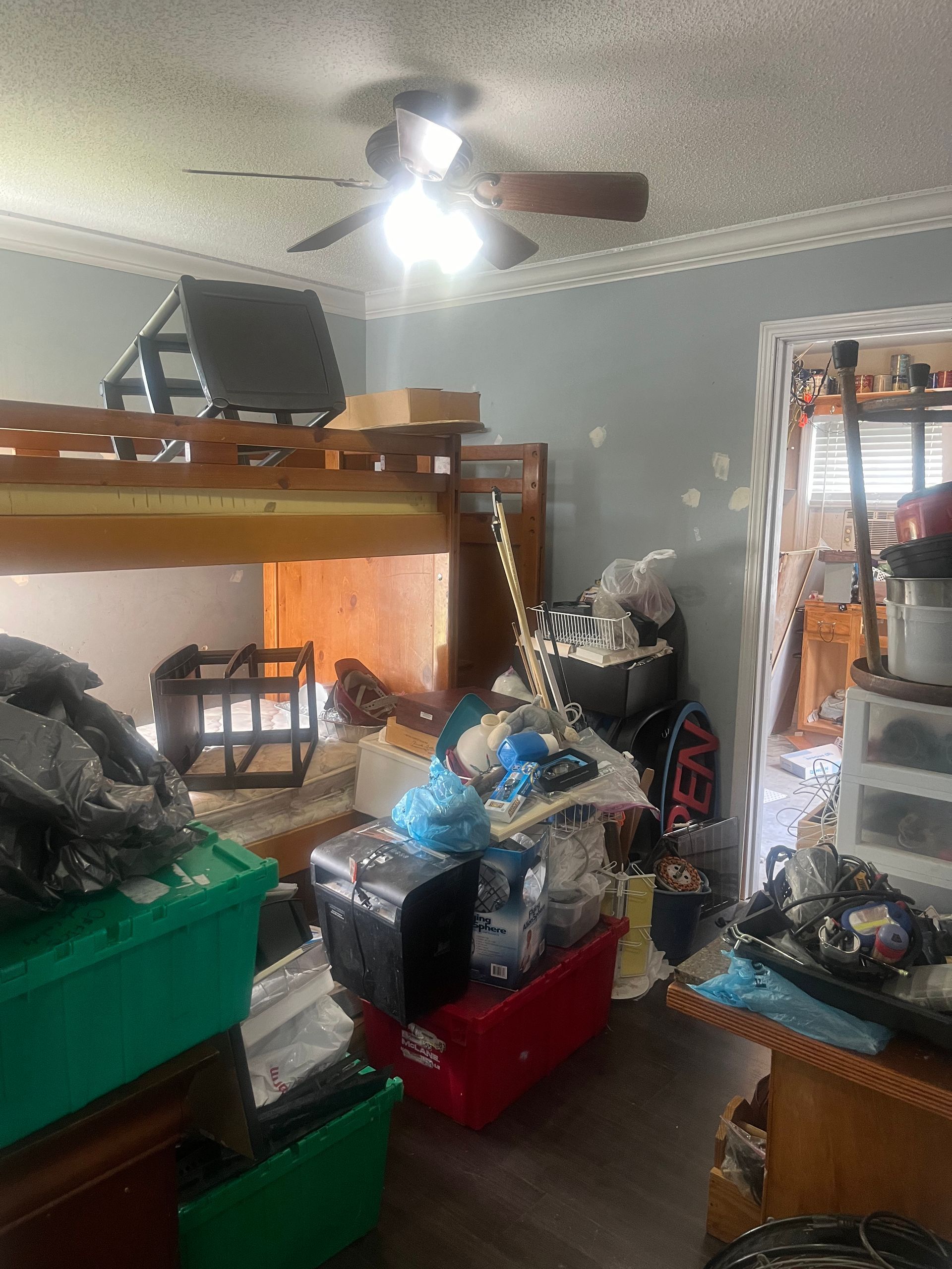 Cluttered room with bunk bed, storage bins, and assorted items. A light shines from the ceiling fan in the center.