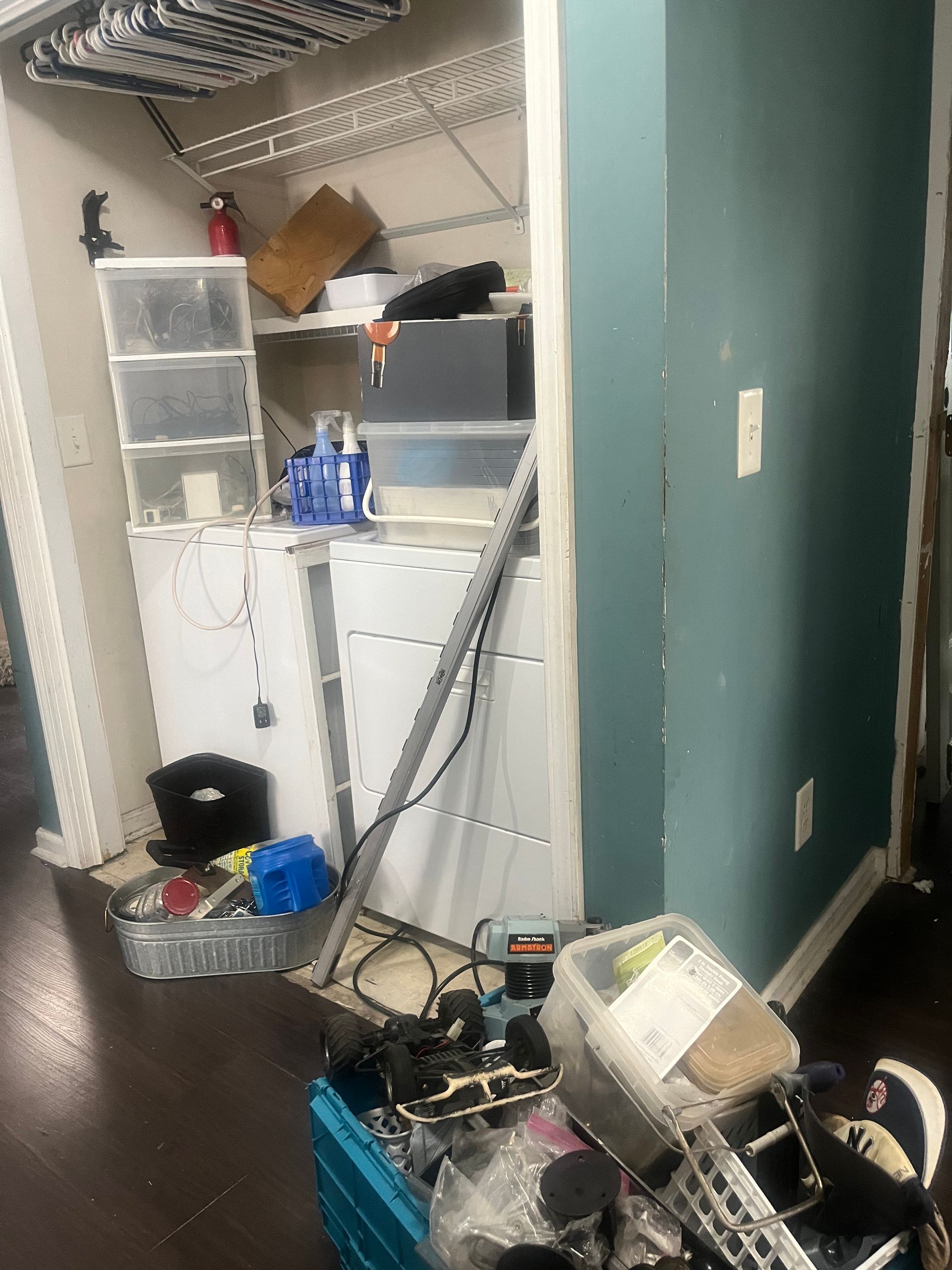 A cluttered room interior with a washing machine, shelves, and various items. Dark wood floor, blue wall.