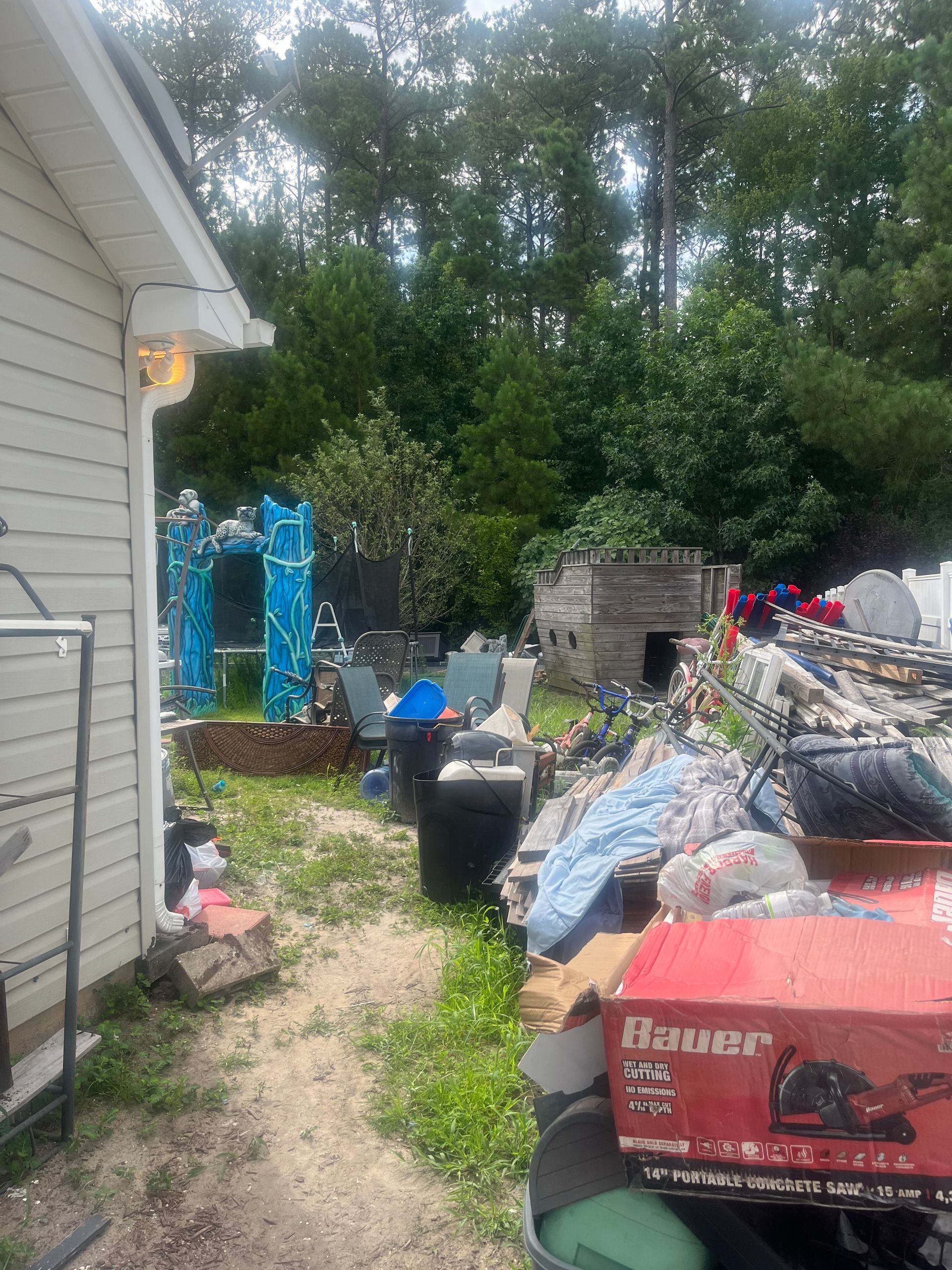 Backyard scene with assorted items. To the left, a white building's corner. Behind the items, trees and blue decorative structures are visible.