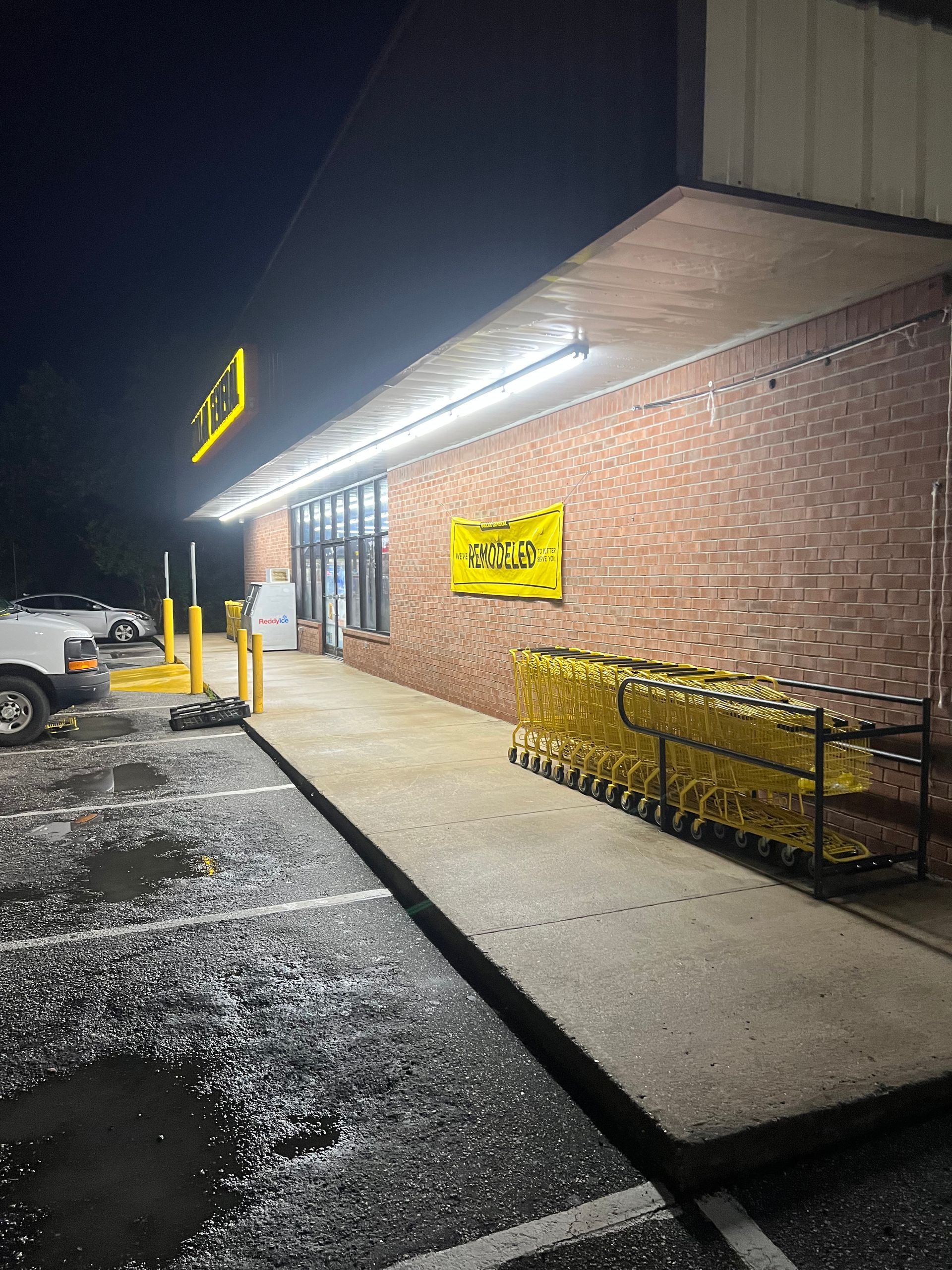 Nighttime view of a Dollar General store entrance with yellow signs and shopping carts. A concrete ramp leads to the glass doors.