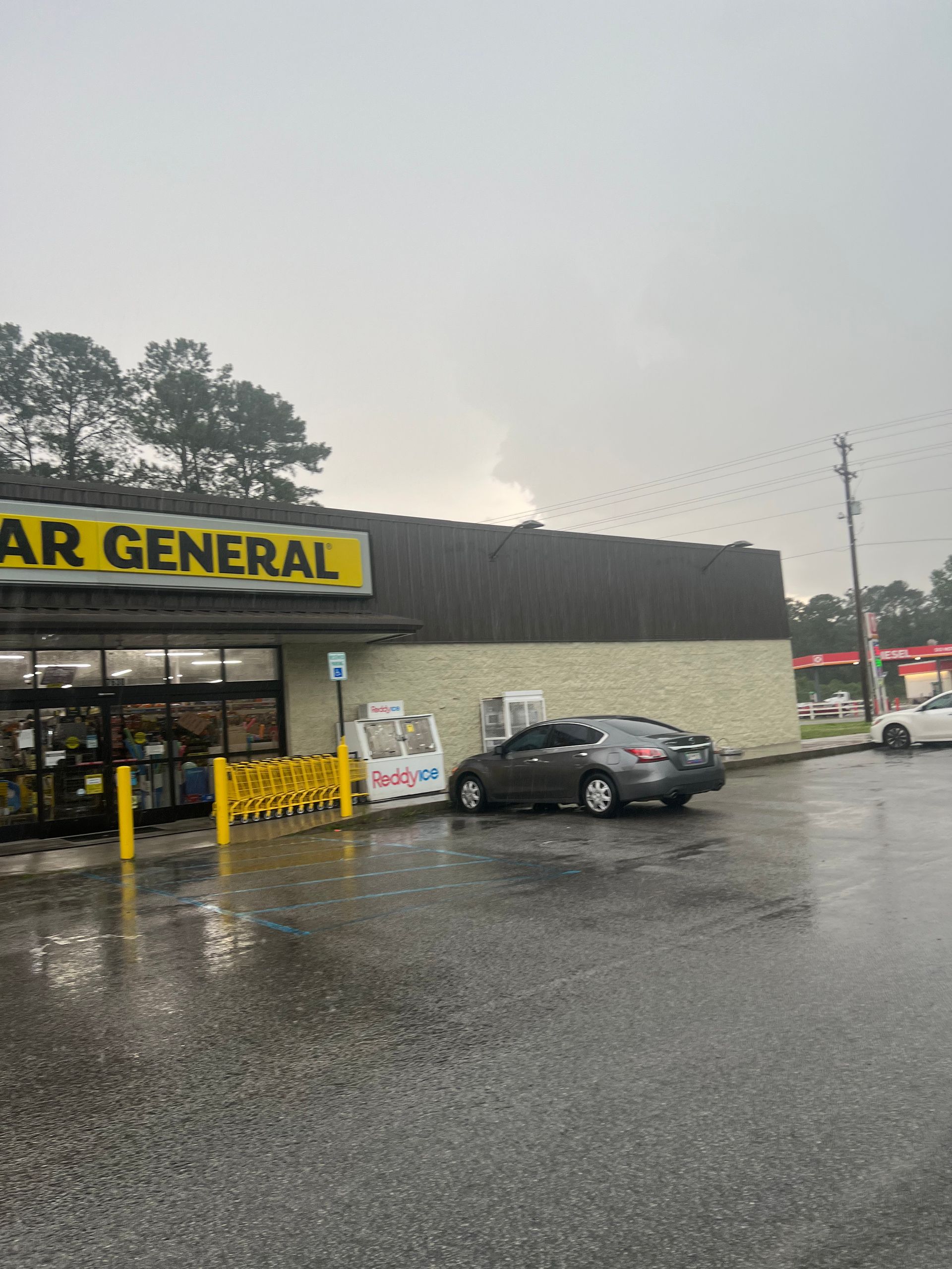 Dollar General store on a rainy day with a gray car parked near the entrance. Yellow sign reads 
