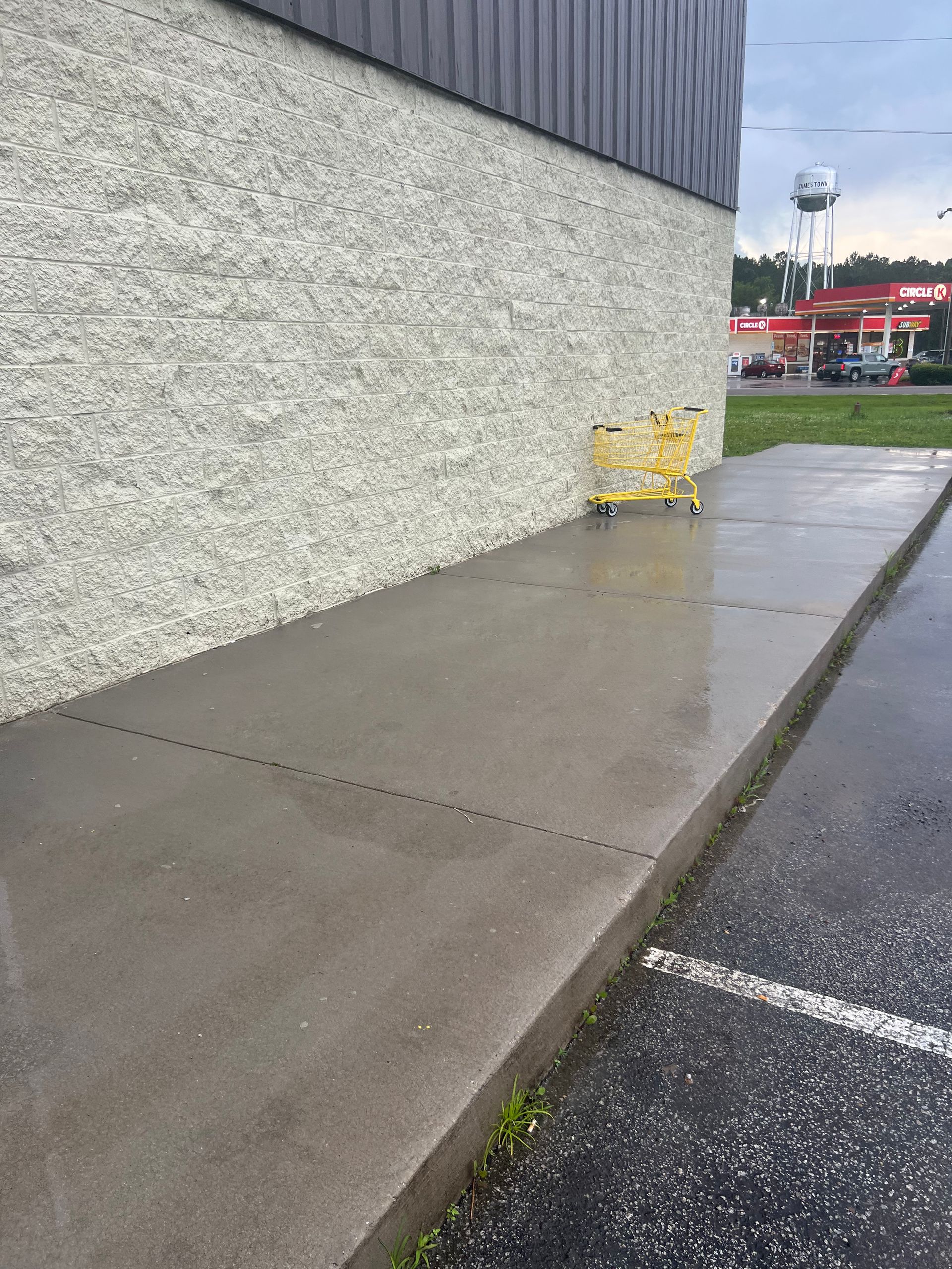 Wet concrete sidewalk next to a textured gray building and a parked shopping cart. In the background are a water tower, cars, and a restaurant.