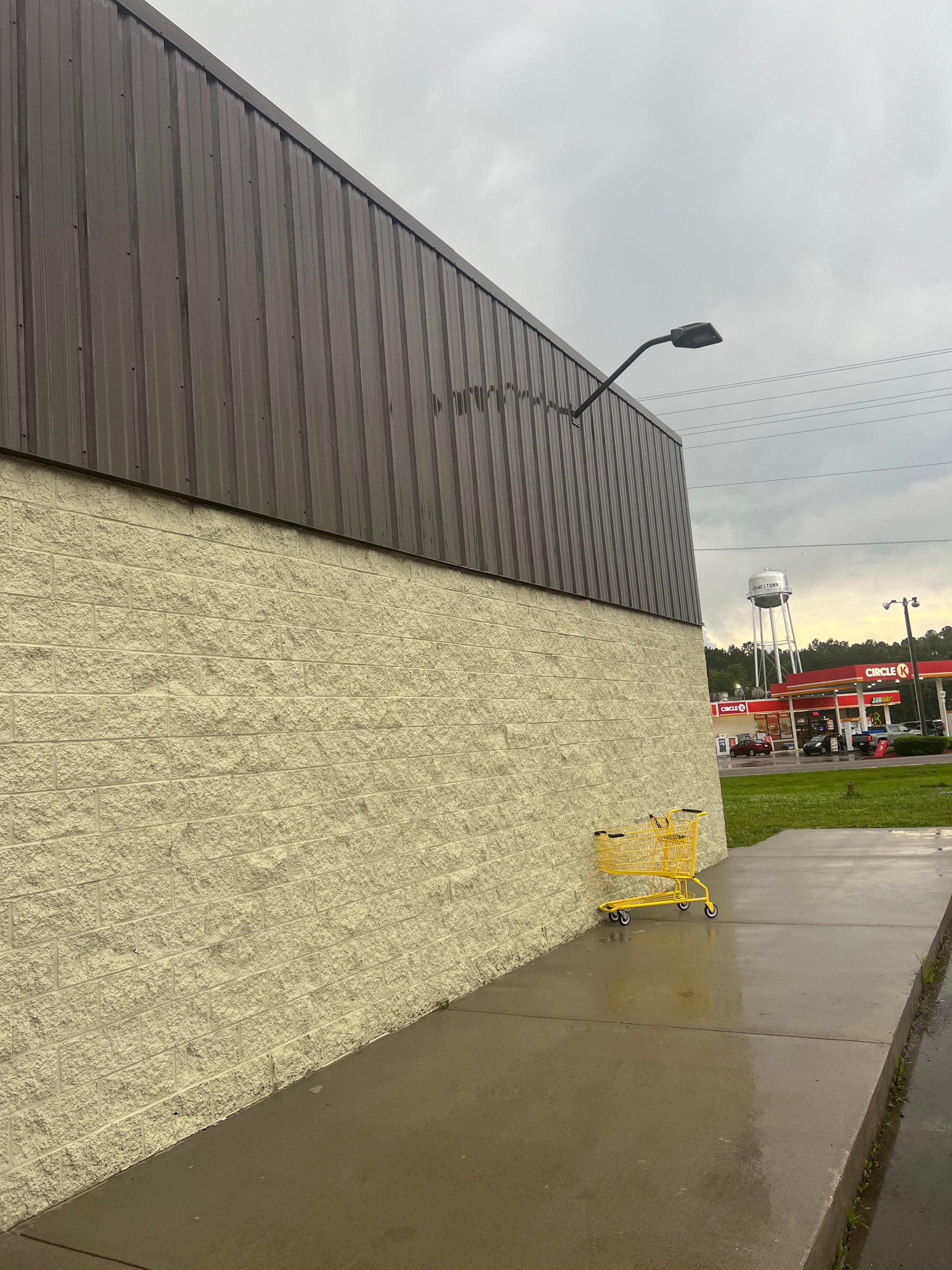 Exterior view of a building with a textured tan lower wall and brown upper wall. A wet sidewalk and yellow shopping carts are in front.