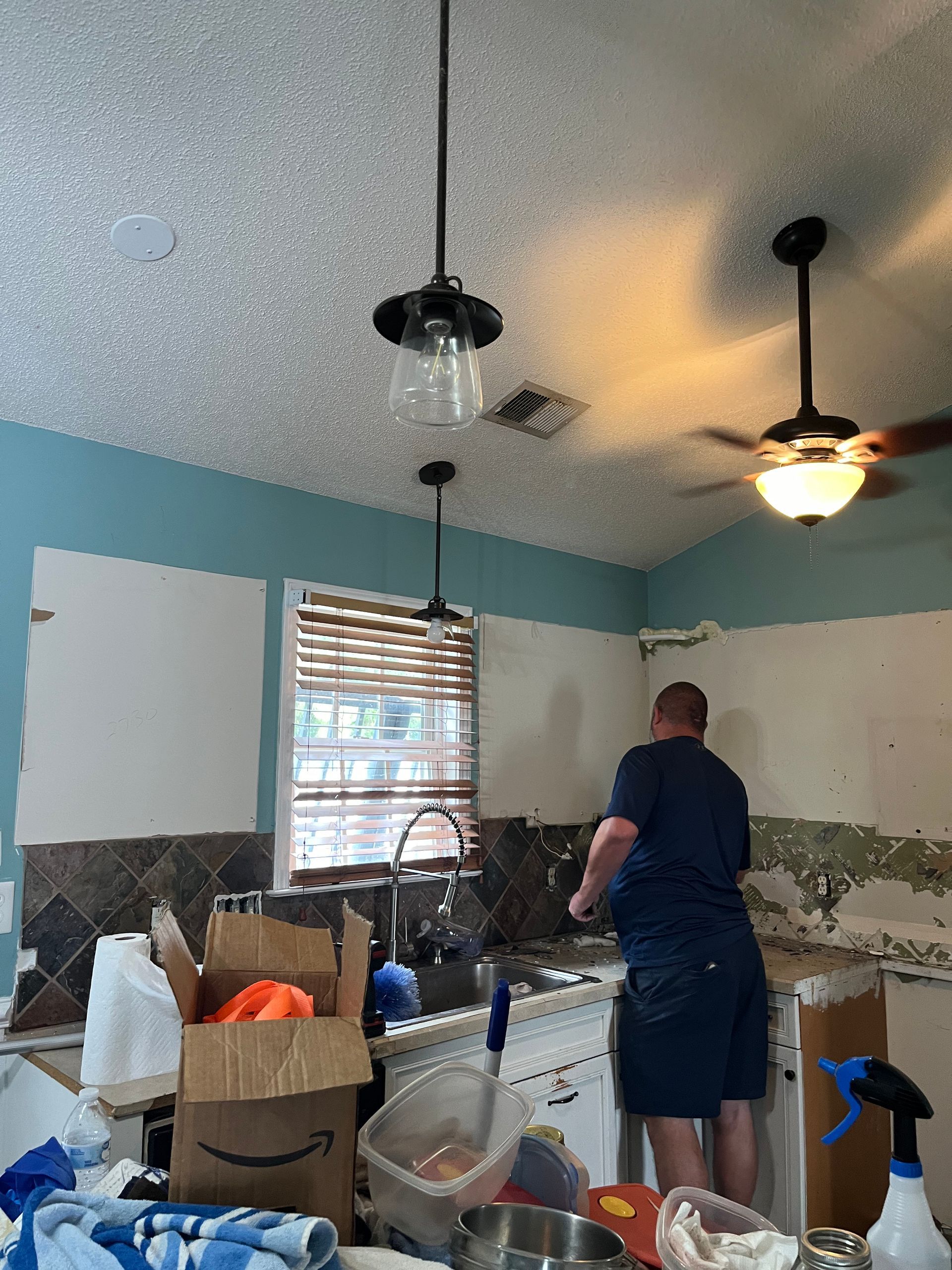 A person works in a kitchen with exposed walls, plumbing, and light fixtures. The ceiling is textured and the walls are blue and white.