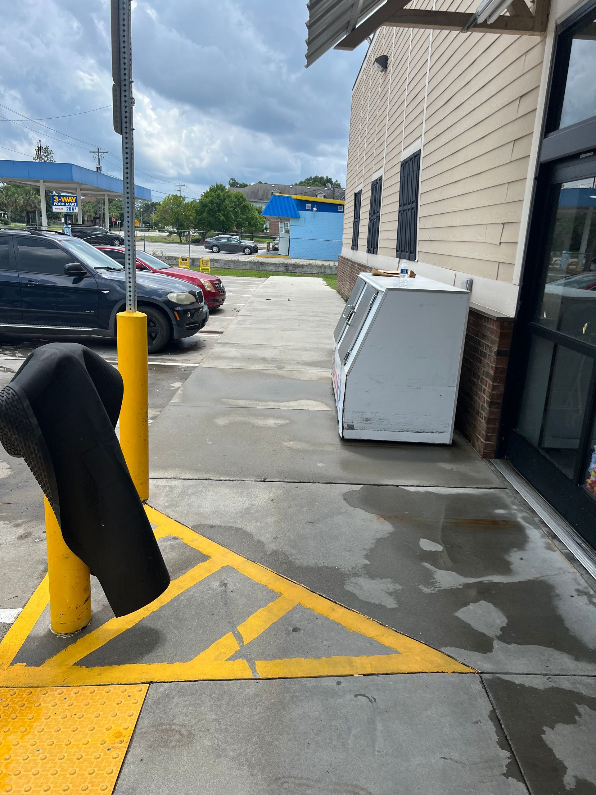 Sidewalk with yellow painted markings, leading to a commercial building entrance. A cooler sits outside. A yellow pole stands in the foreground with a black item hanging on it.