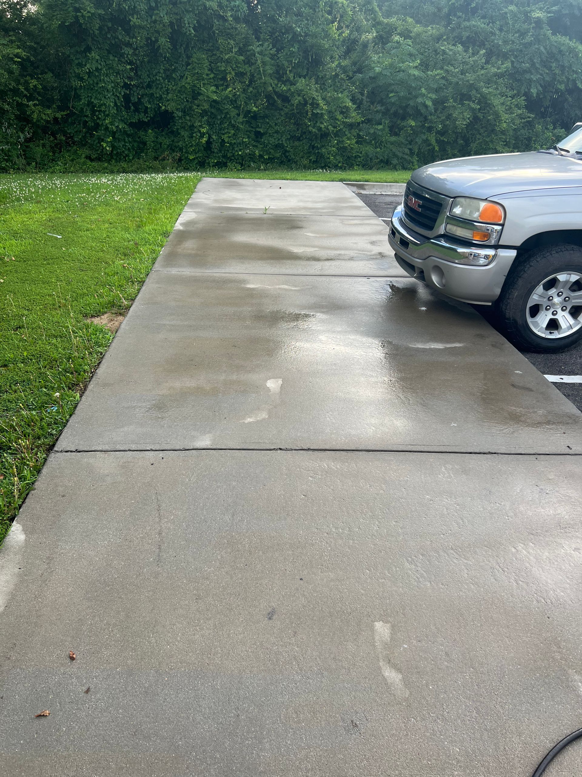 A silver truck parked on a wet, gray concrete driveway next to grass and trees.