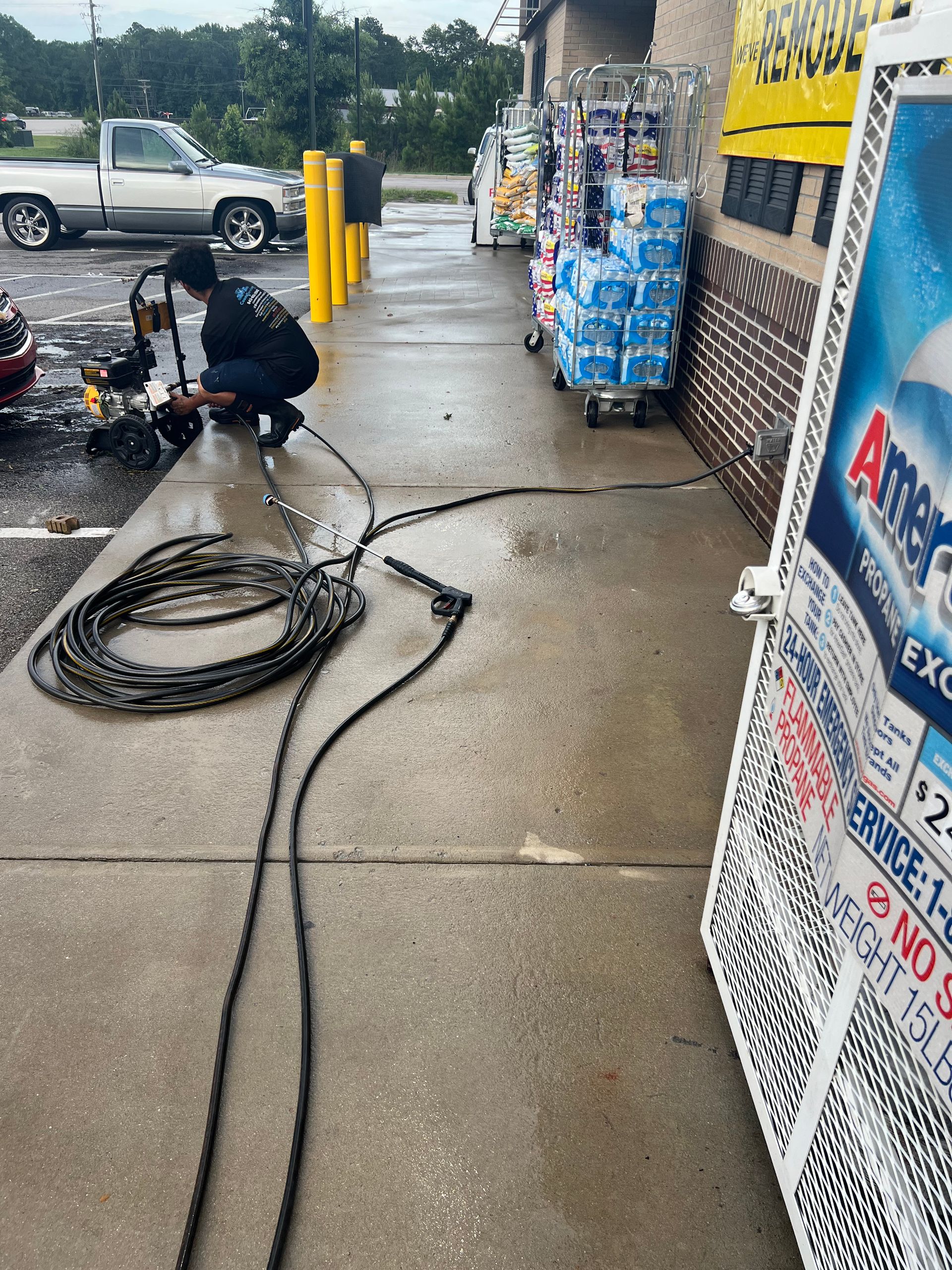 Person power washing a sidewalk outside a store. Wet concrete and equipment visible, including a truck parked nearby.