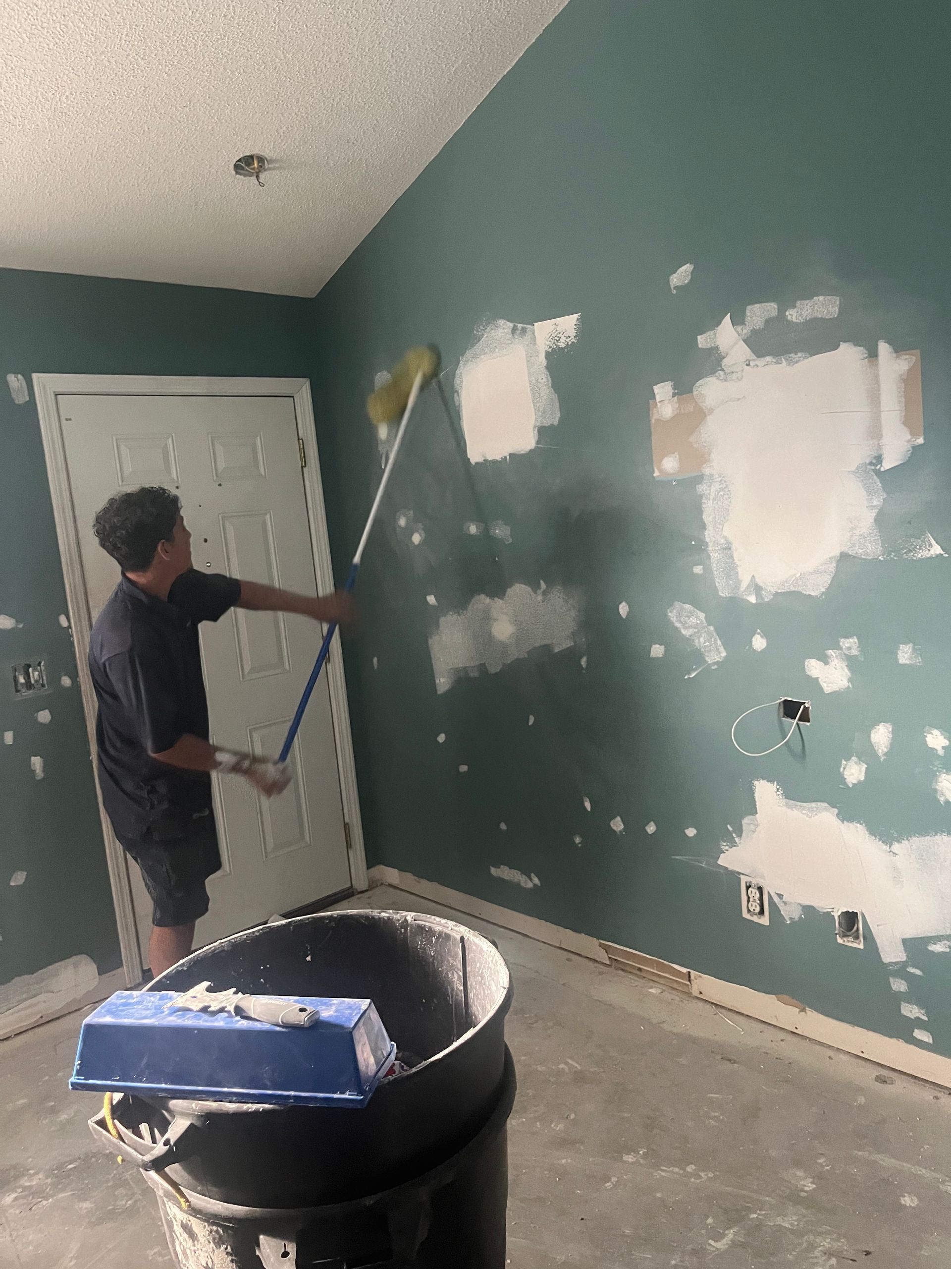 A person in a dark shirt and shorts applies drywall mud to a teal wall. They are using a long-handled applicator in a room with a door and a garbage can.