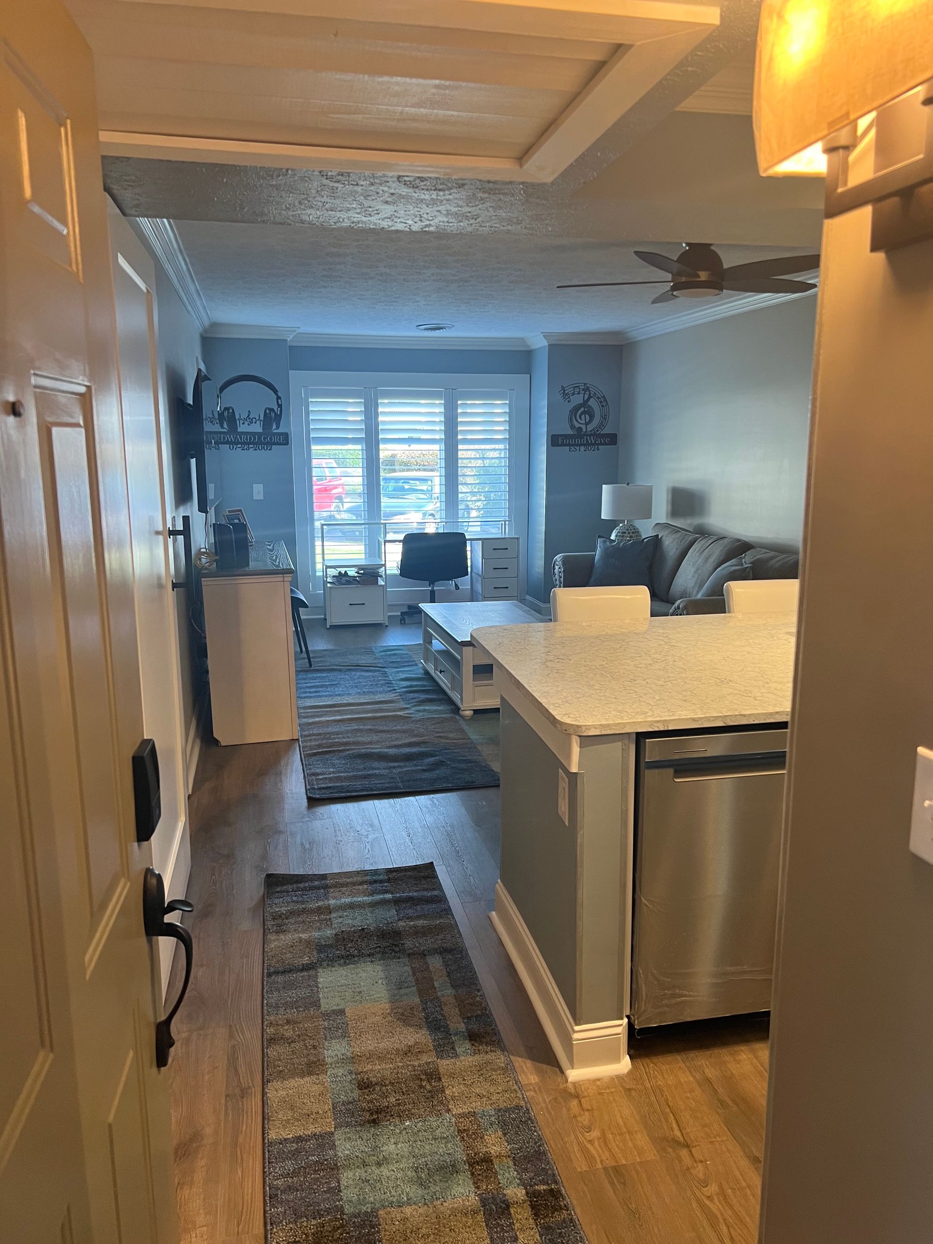 Entryway view into a modern living space with a kitchen island. Light-colored wooden floors, a blue rug, and a window with bright daylight are visible.