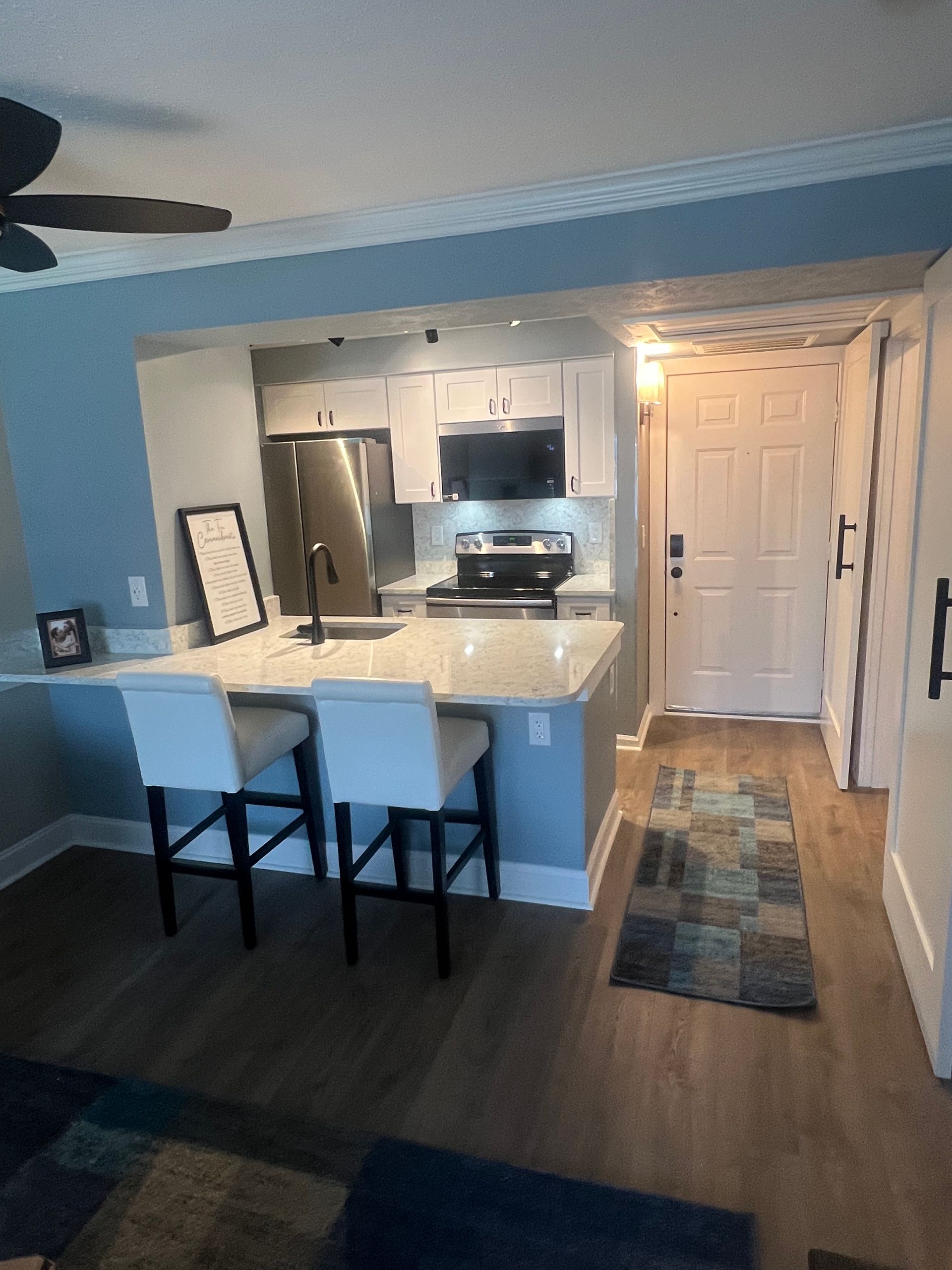 A modern kitchen with white cabinets, a breakfast bar with stools, and a stainless steel refrigerator. A blue rug leads to a white door.