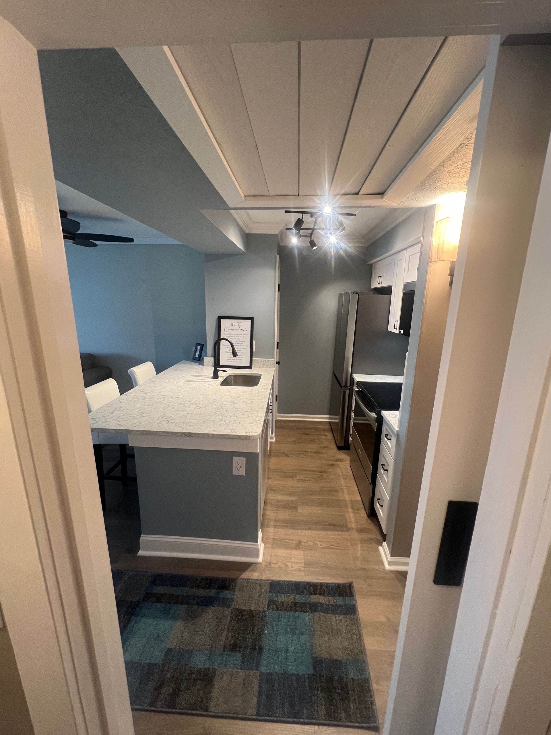 Looking down a hallway into a kitchen and living area with blue and gray accents, wood-look flooring, and a white countertop.
