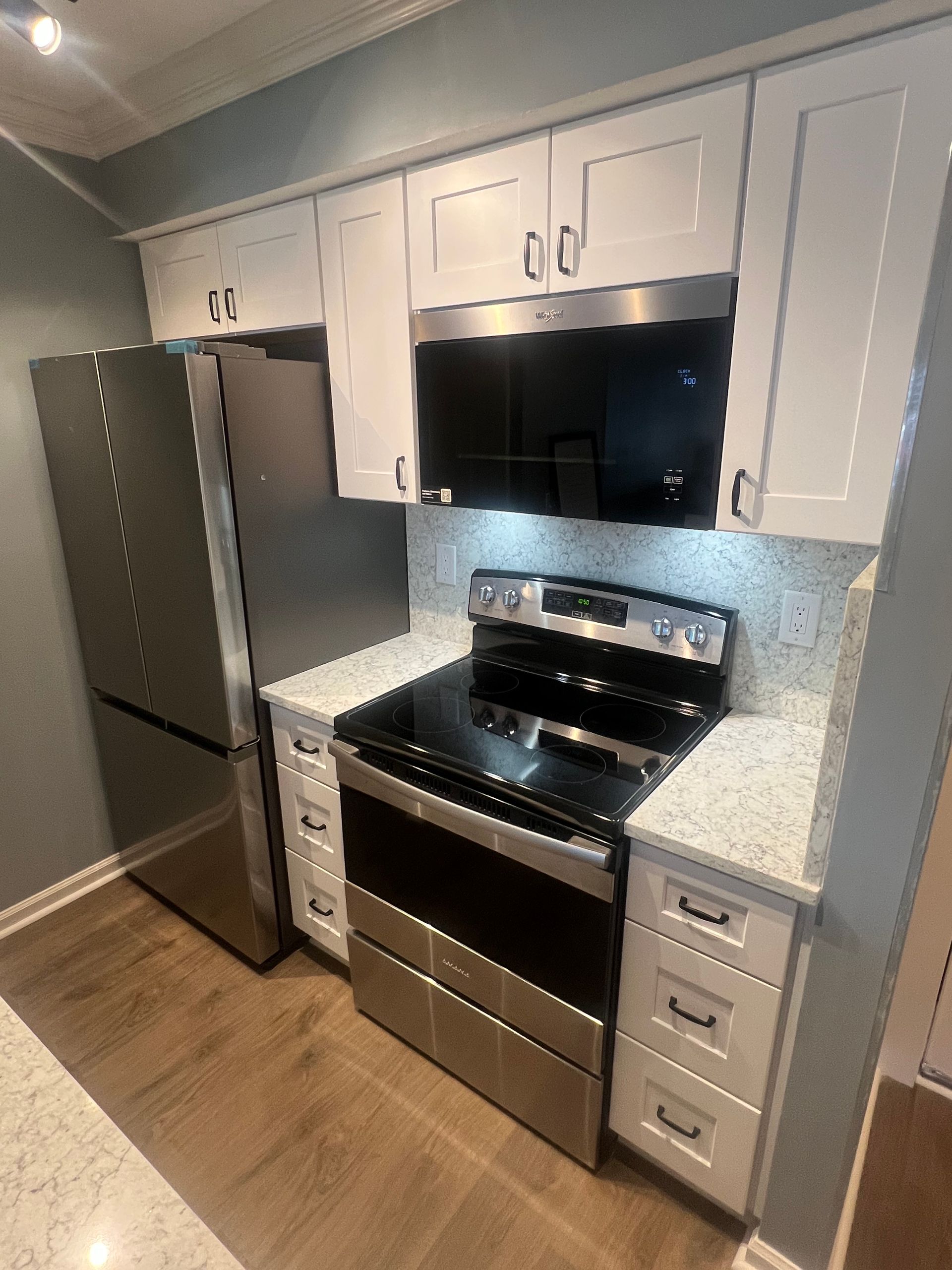 Modern kitchen with white cabinets, stainless steel appliances (refrigerator, range, microwave), and light countertops. Wooden floors.