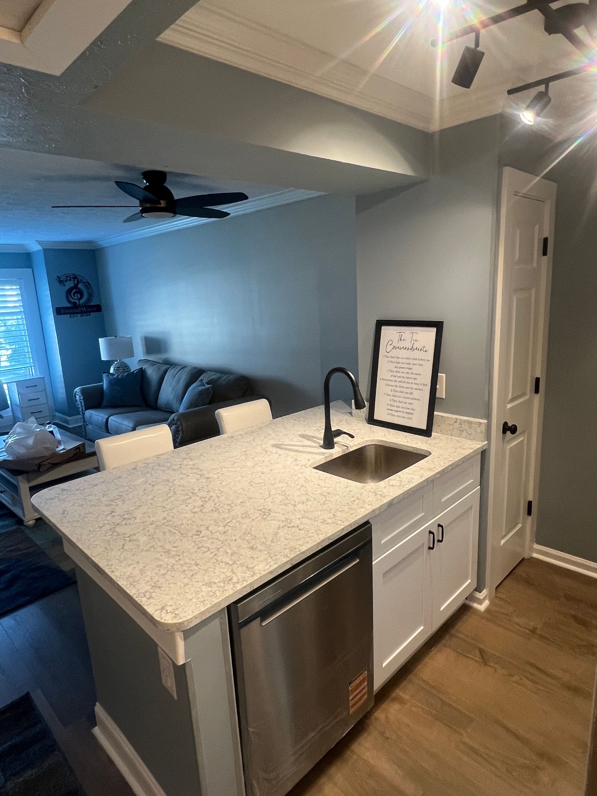 Small kitchen island with a sink and dishwasher. A framed menu is on the countertop. The room has a blue-gray wall and wood-look floors.