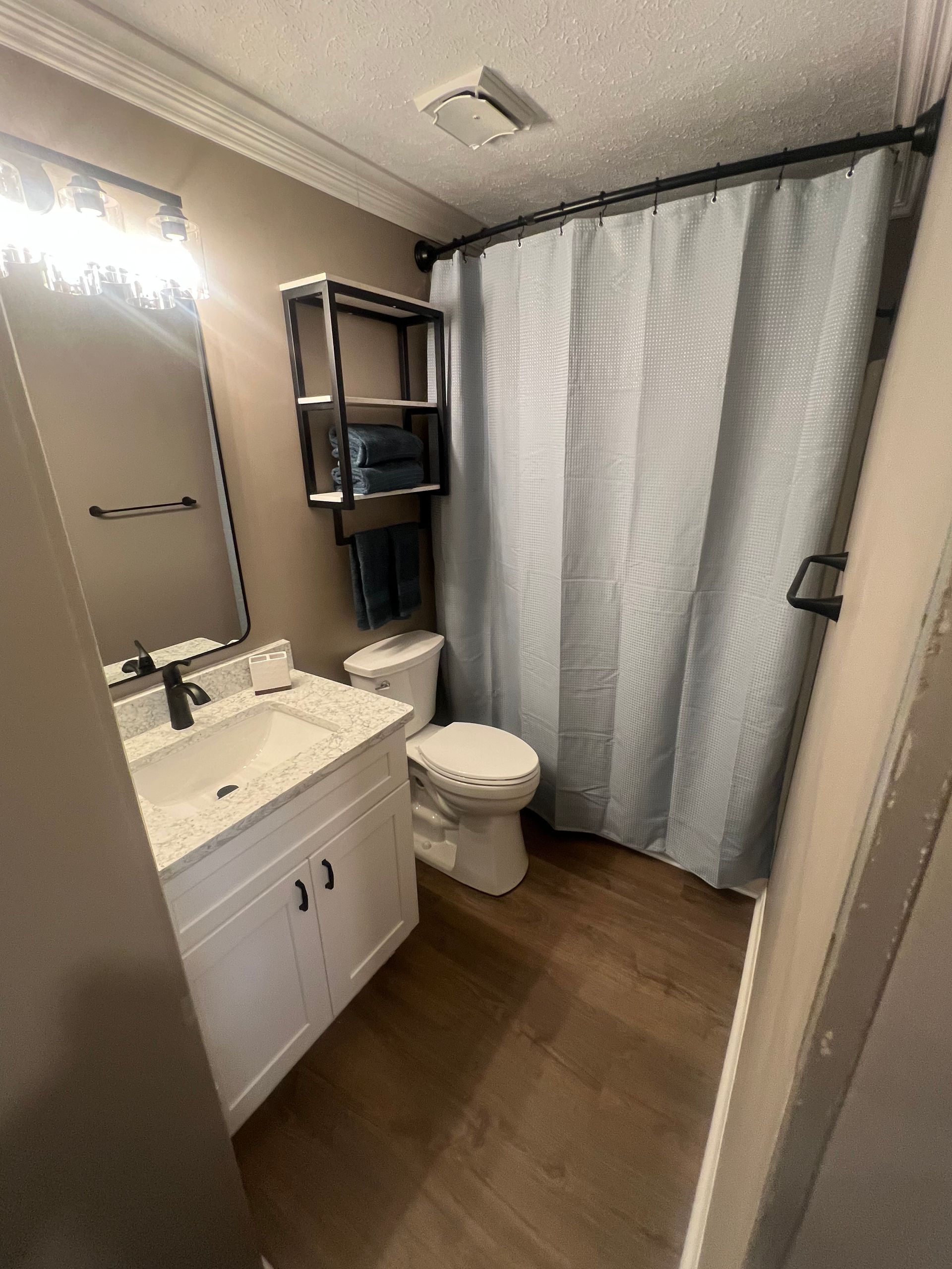 Small bathroom with a white vanity, toilet, and light blue shower curtain.  Black shelf with towels and a window.  Wooden floor, tan walls.