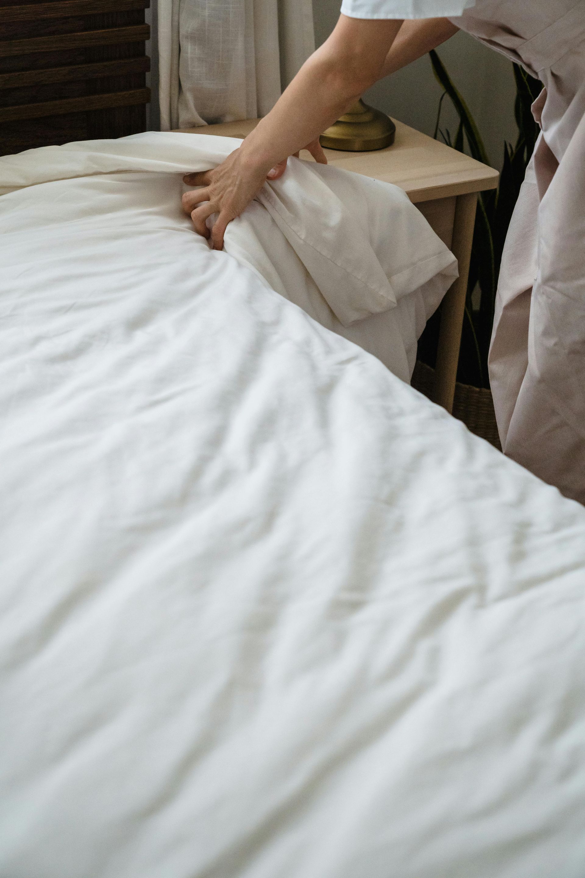 Person making a bed, smoothing white sheets. They're standing near a light-colored wooden nightstand.