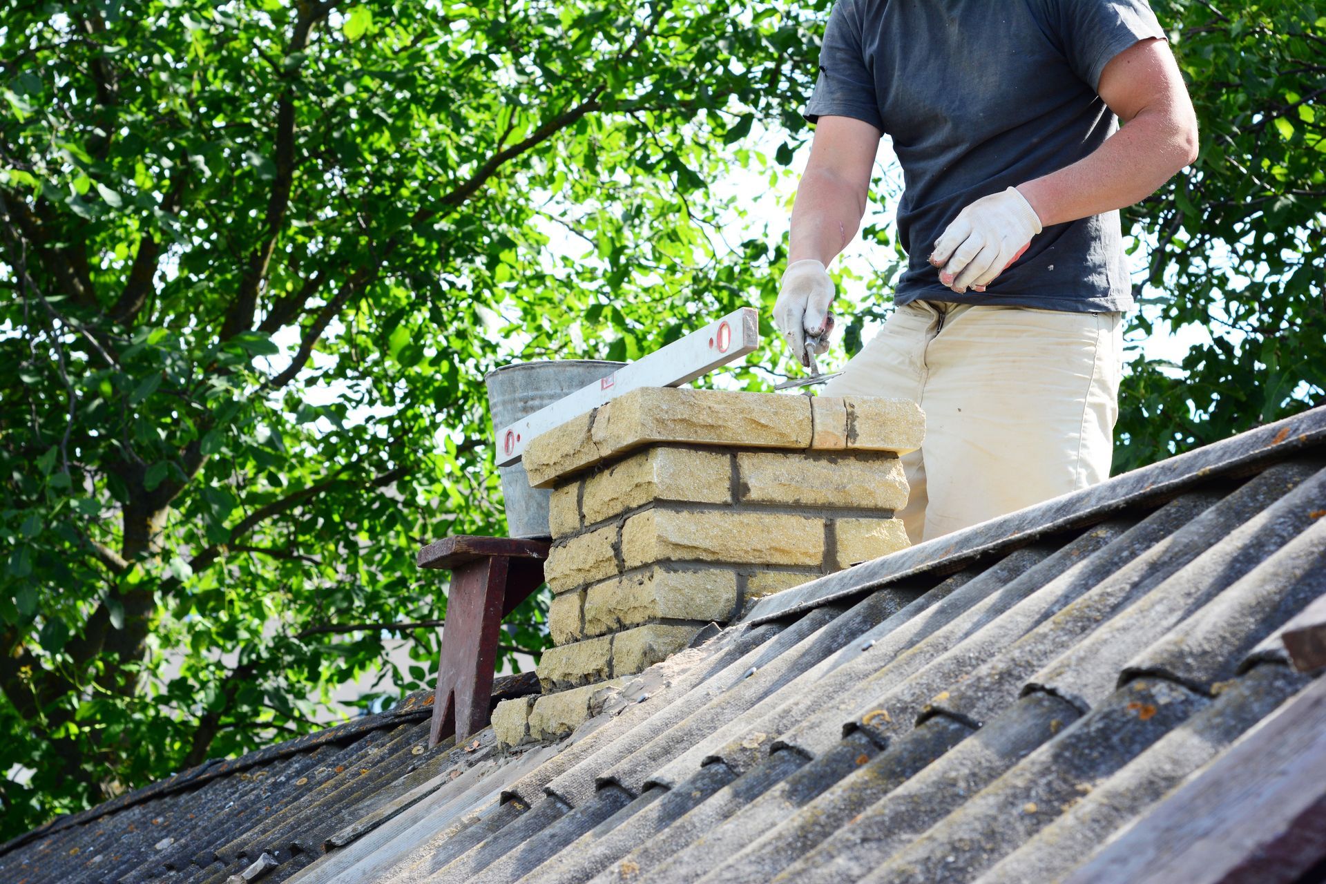 Person on a roof near a brick chimney, wearing gloves, bucket and tools. Green trees in background.