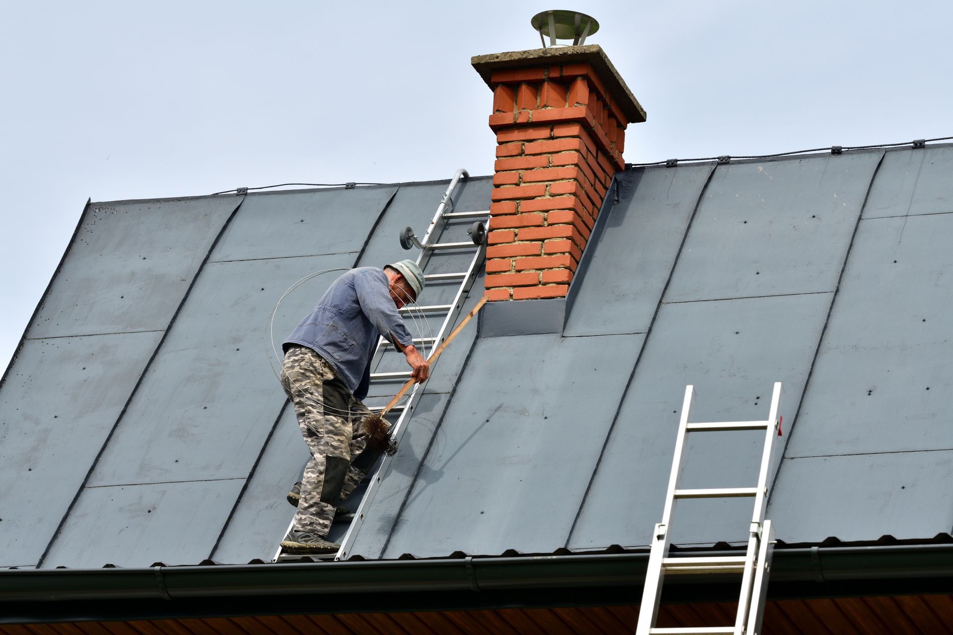 Man on a roof climbs a ladder to a chimney. Gray roof, brick chimney, clear sky.
