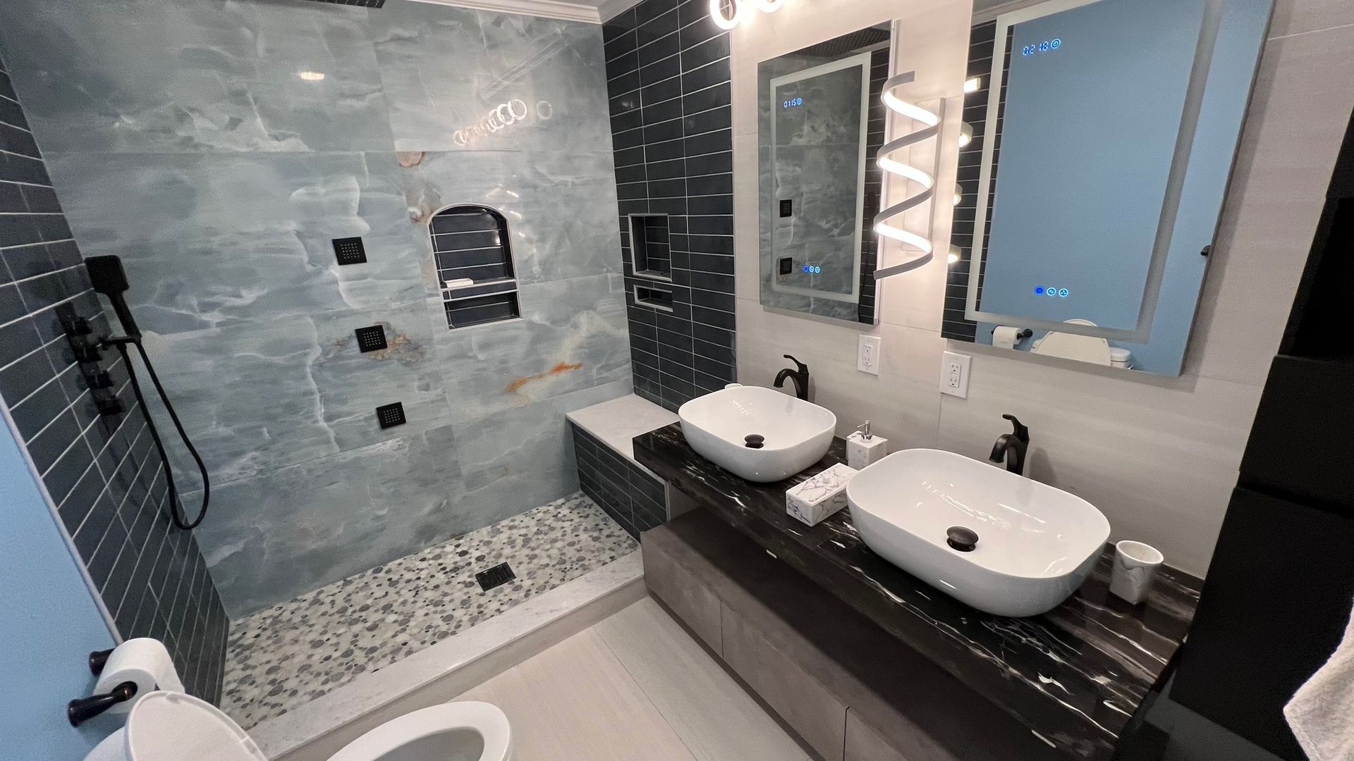 Modern bathroom with a dual-sink vanity, backlit mirrors, and a walk-in shower featuring blue tile and pebble flooring.