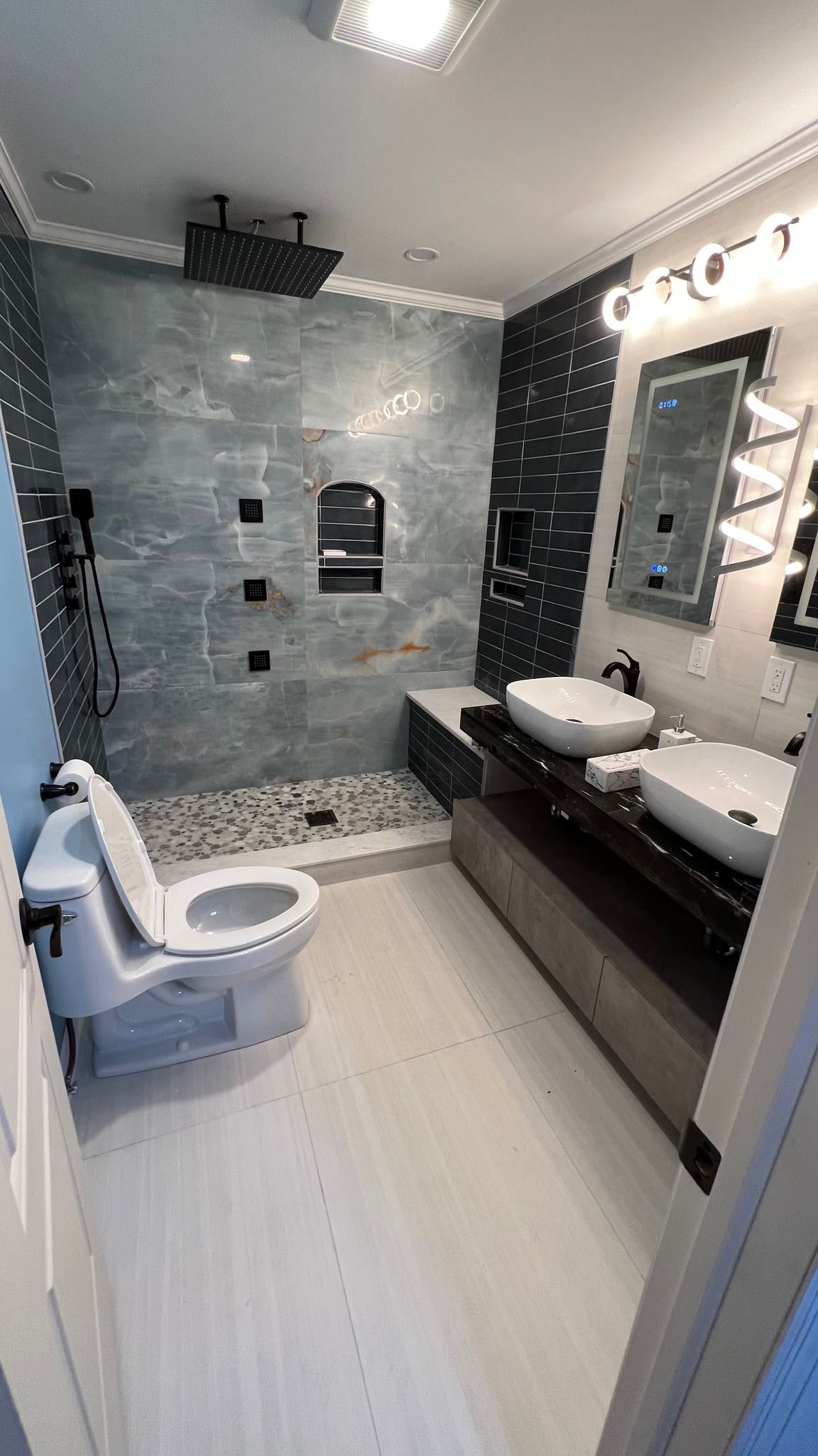 Modern bathroom with a walk-in shower, double vanity, and toilet, featuring gray stone tiles and a dark vanity cabinet.