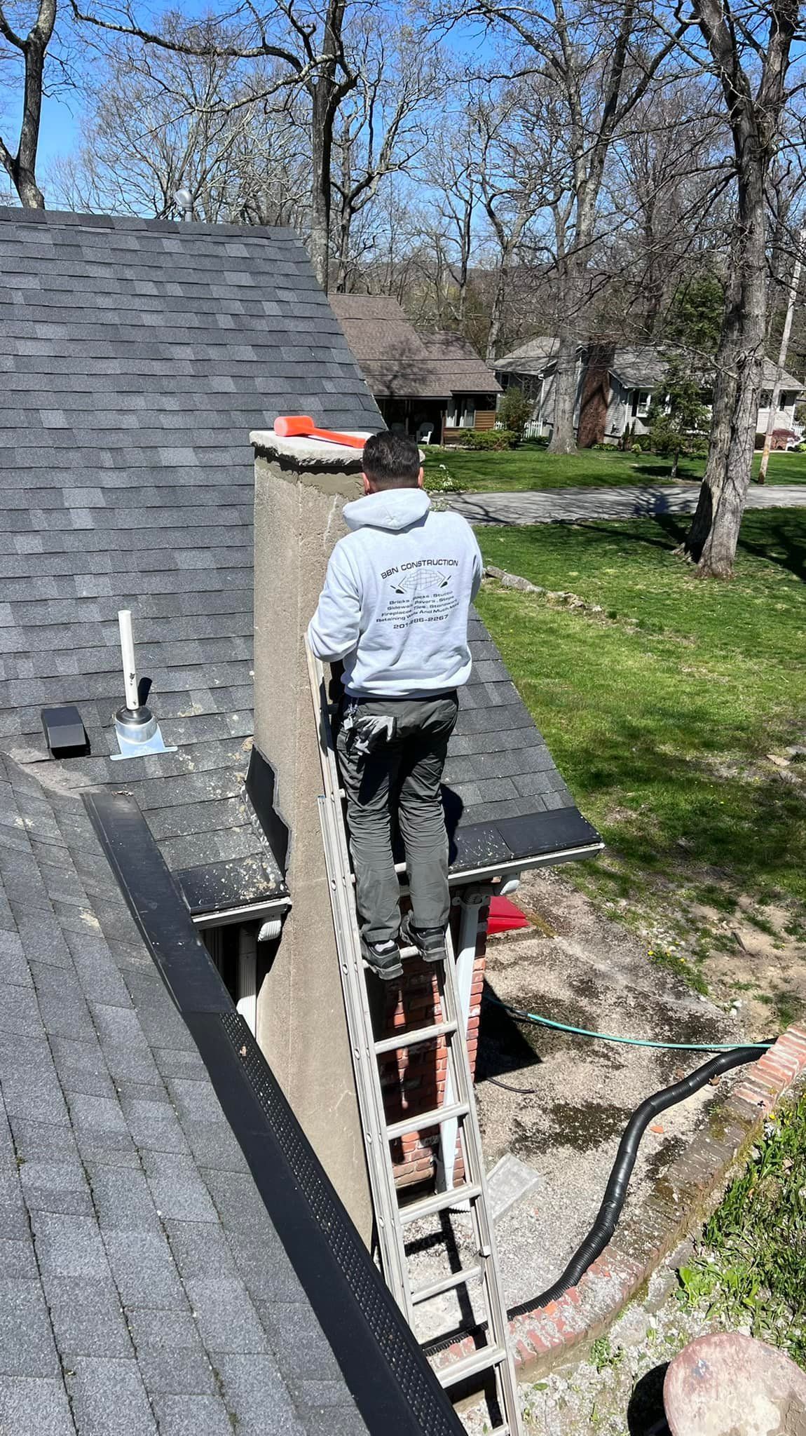 Roofer on a roof, inspecting a brick chimney in a neighborhood with homes and trees in the background.