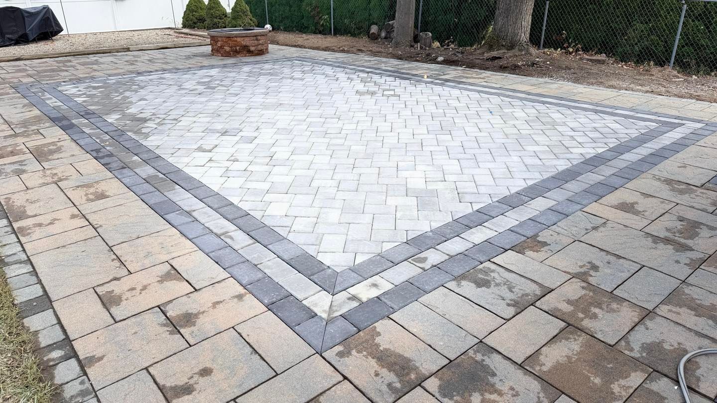 A rectangular patio made of light gray herringbone pavers with a dark gray border, set in an outdoor yard.