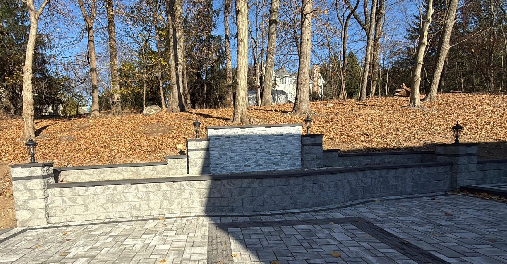 Stone retaining wall and paved area, with trees and fallen leaves in the background.