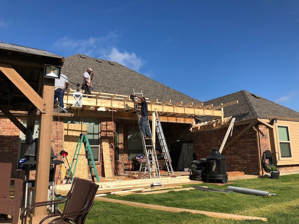 Construction workers building a covered patio extension onto a brick home on a sunny day. They are working on the wooden frame.