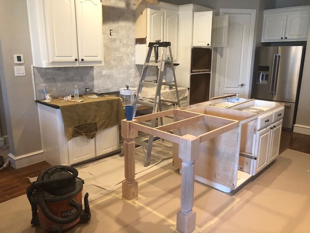 Kitchen renovation in progress: white cabinets, unfinished island with wooden supports, ladder, stainless steel refrigerator, beige flooring.