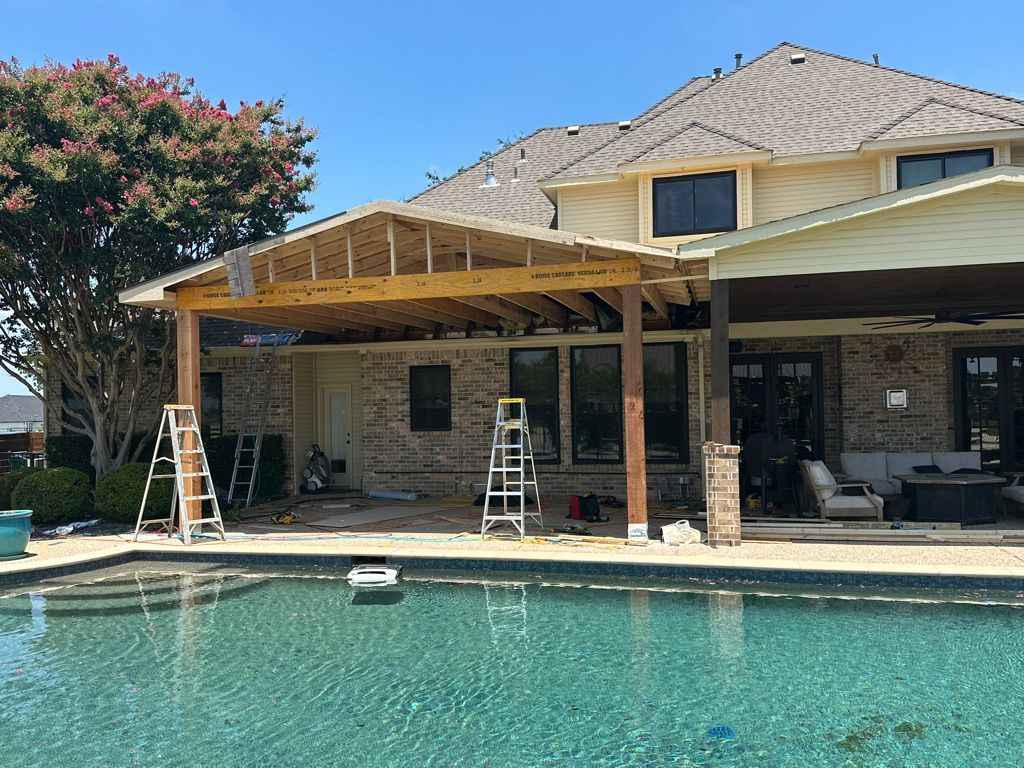 Construction in progress on a covered patio addition next to a swimming pool. Partially built roof with visible wood beams, near a brick home.