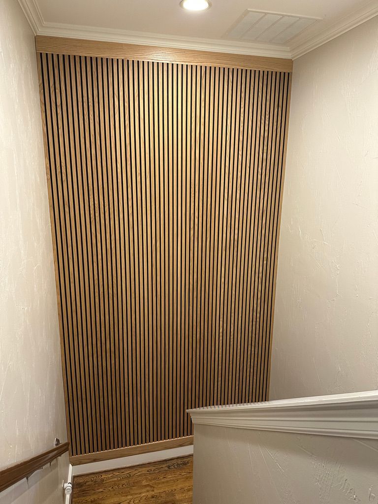 Wooden slat wall with a wavy pattern, framed by a light-colored molding. The wall is in a stairwell with light-colored walls.