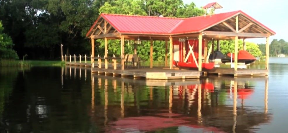 Red-roofed boat dock over calm lake, with patio chairs and covered boat storage.