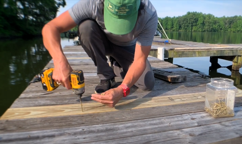 A person repairing dock with drill near water and trees