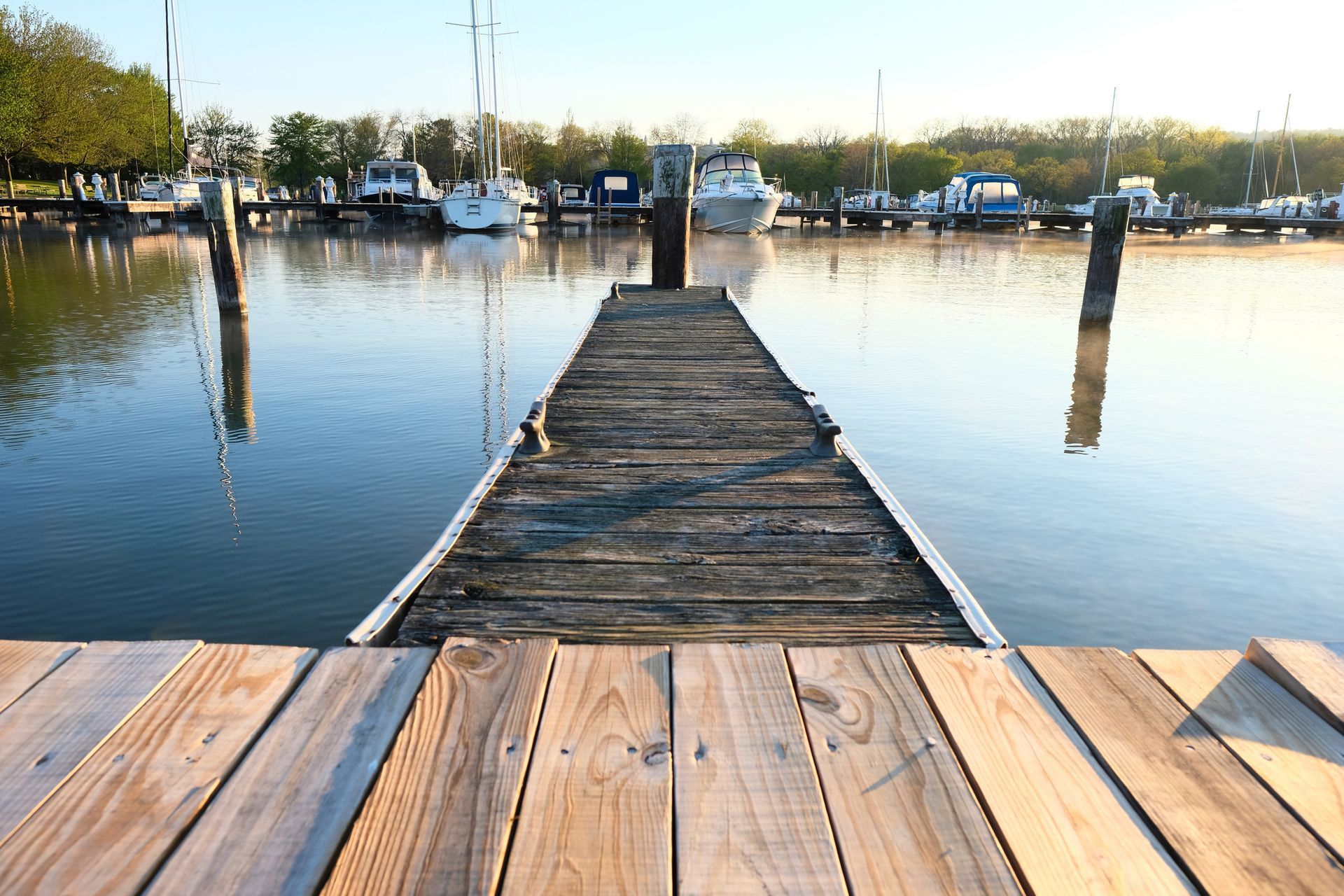 A wooden dock over calm water at sunrise with boats and trees in the background