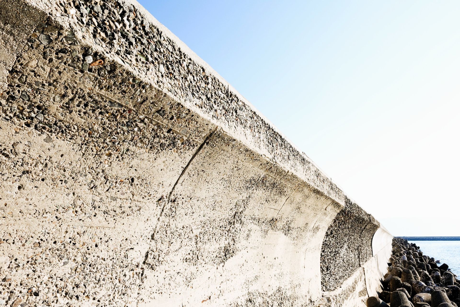 Curving sea wall made of concrete with pebbles against a sunny sky.