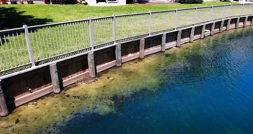 A waterfront view with a wooden barrier, clear water, green grass, and a fence along a seawall.