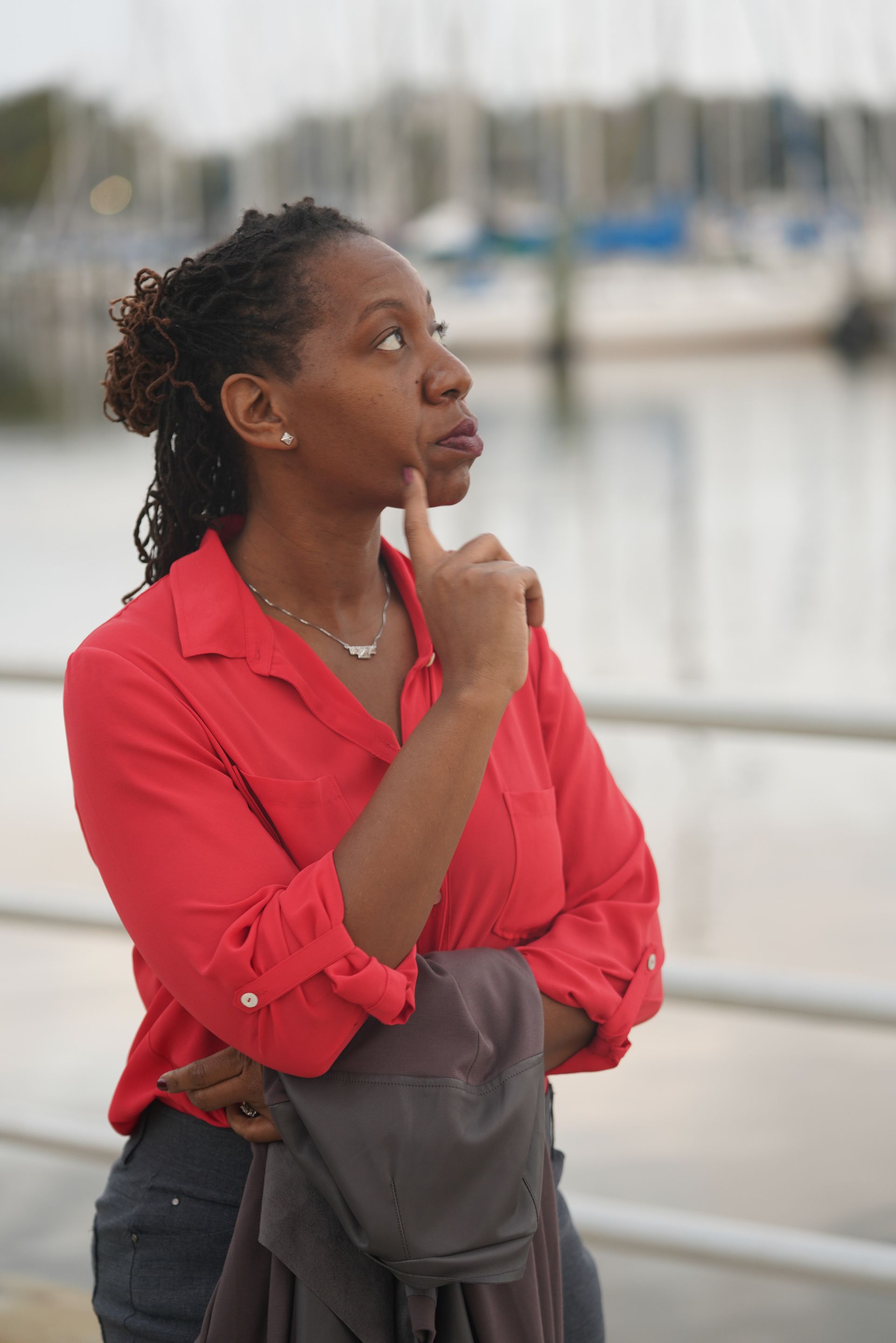 Woman with locs wearing a red shirt, looking up thoughtfully at a marina; she has her finger on her chin.