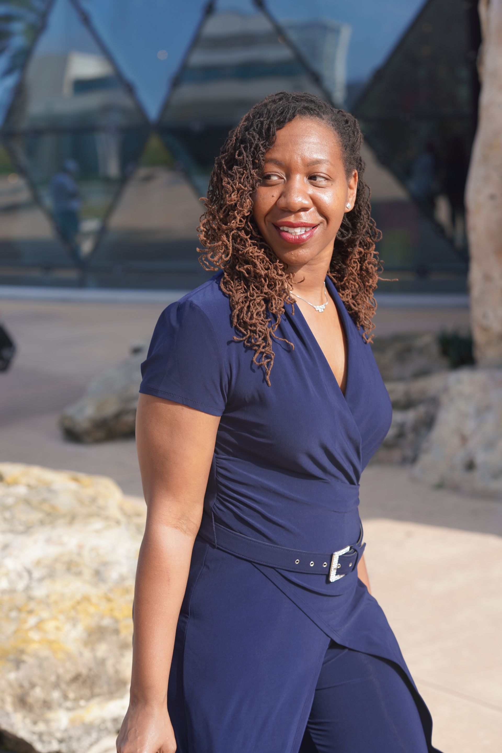 Woman in a dark blue jumpsuit stands outside with a building featuring geometric glass panels in the background.