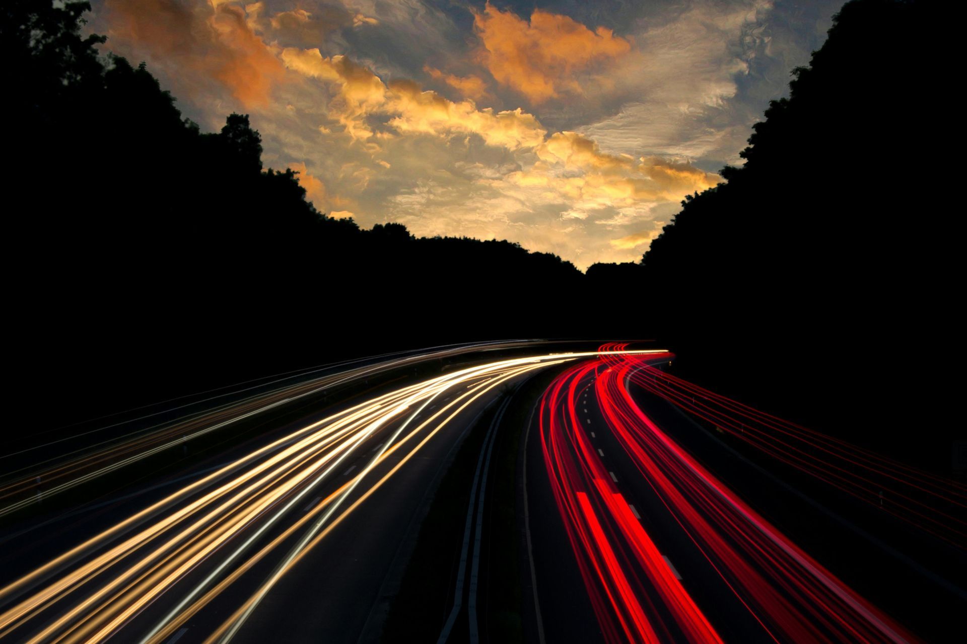 Highway at dusk, with streaking light trails of vehicles in red and white against dark trees and a sunset sky.
