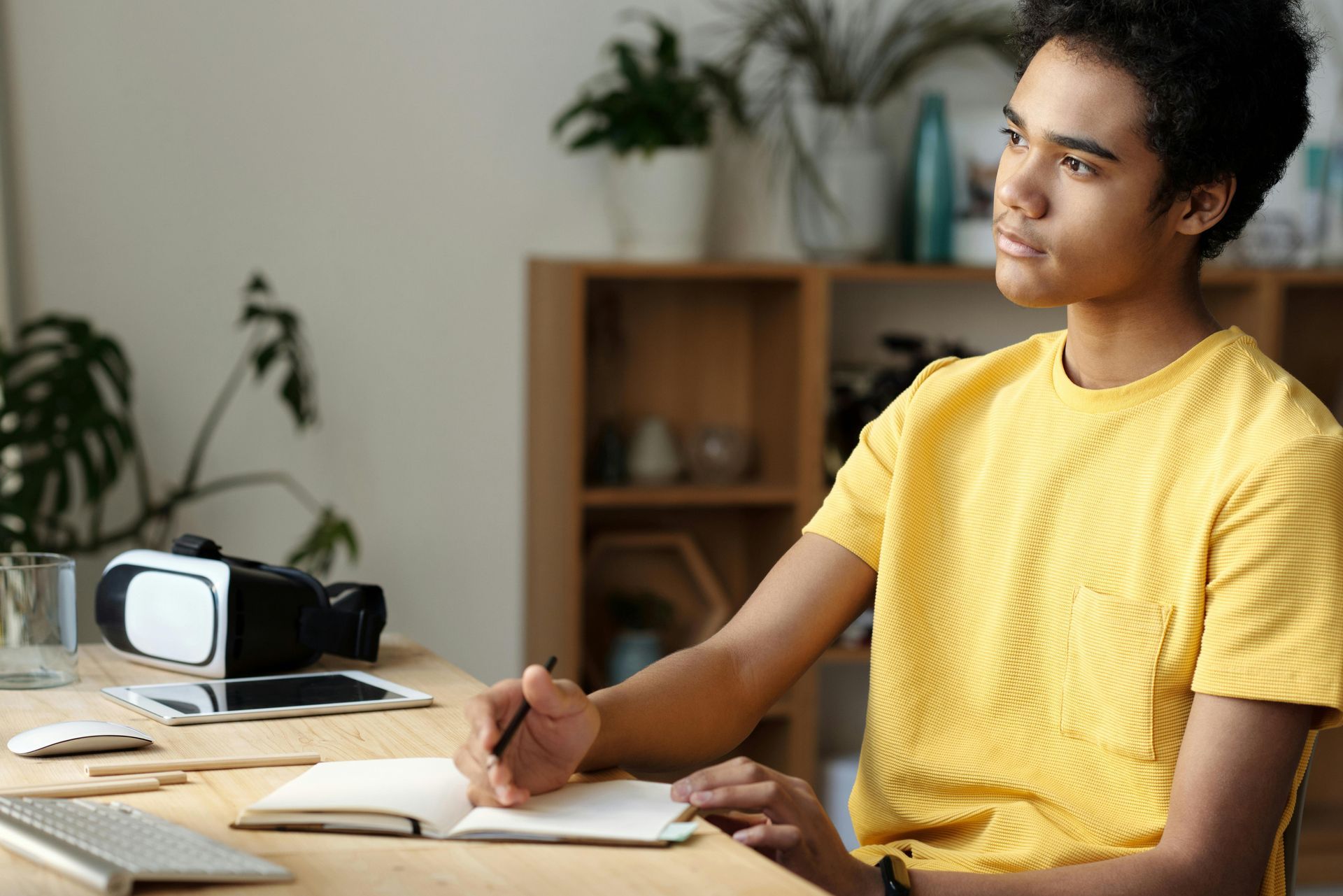 Teenager in yellow shirt, pensively looking up while writing in a notebook at a desk with VR headset and tablet.