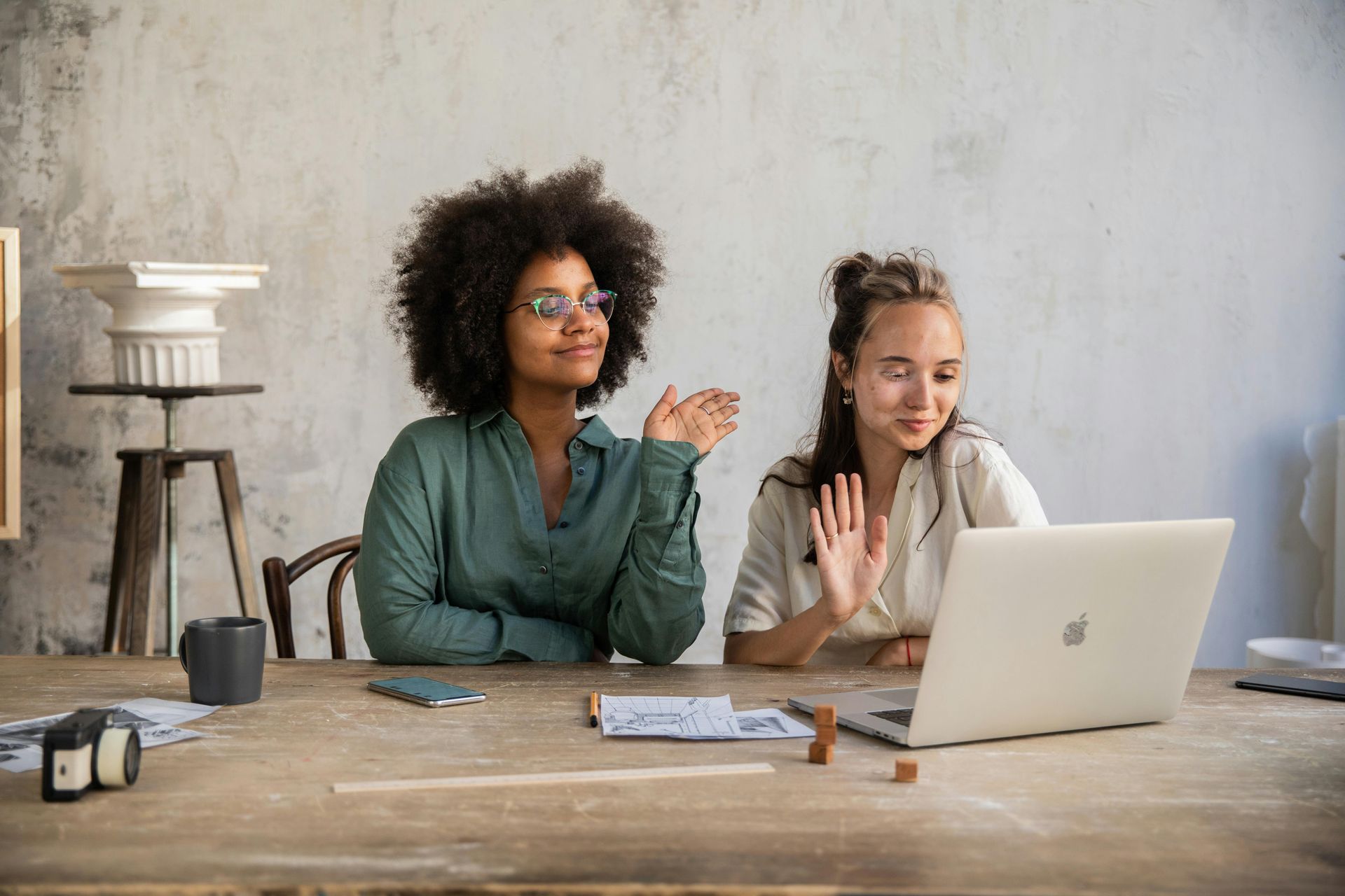Two women are sitting at a table participating in meeting on a laptop computer.