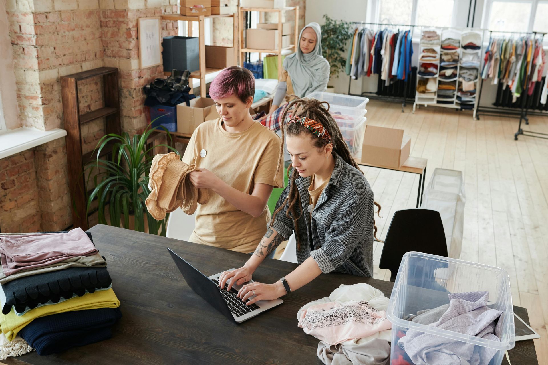 Three people organizing clothing in a brightly lit room. One types on a laptop, one examines clothes, and one stands in the background.