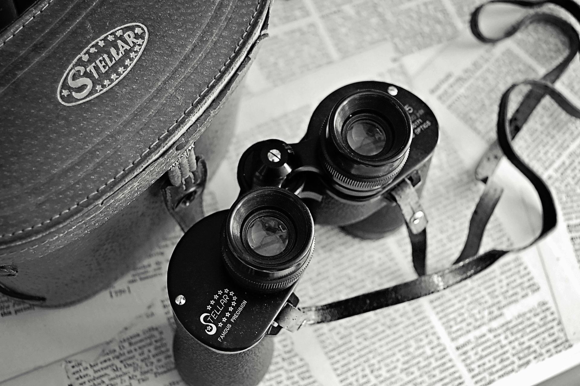 Black and white photograph of binoculars with a leather case, sitting on top of newspaper clippings.