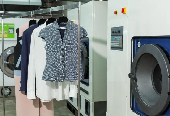 Clothes on hangers in a dry cleaning facility next to industrial washing machines.