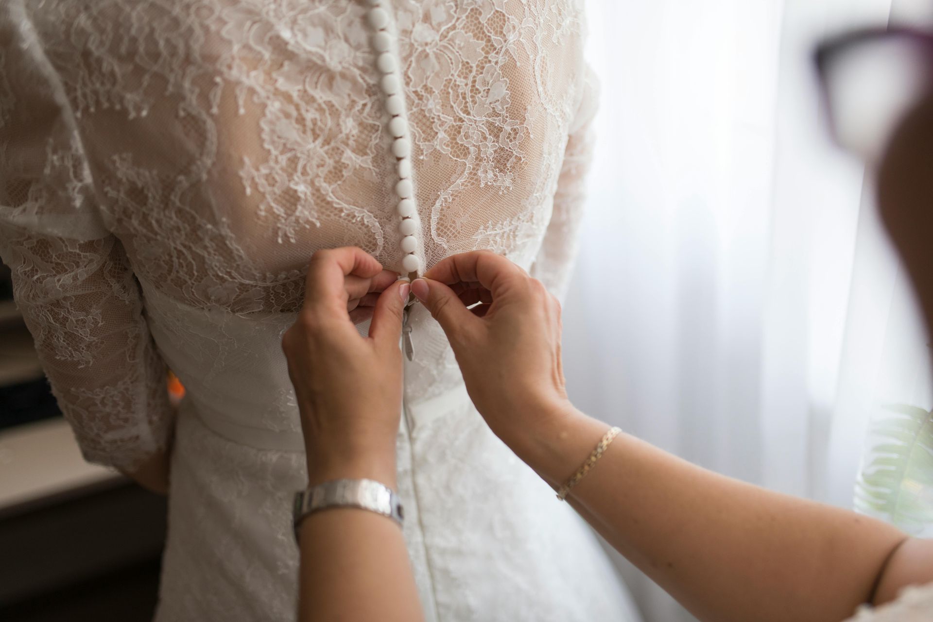 Woman's wedding dress back being buttoned; lace detail, white fabric, hands.