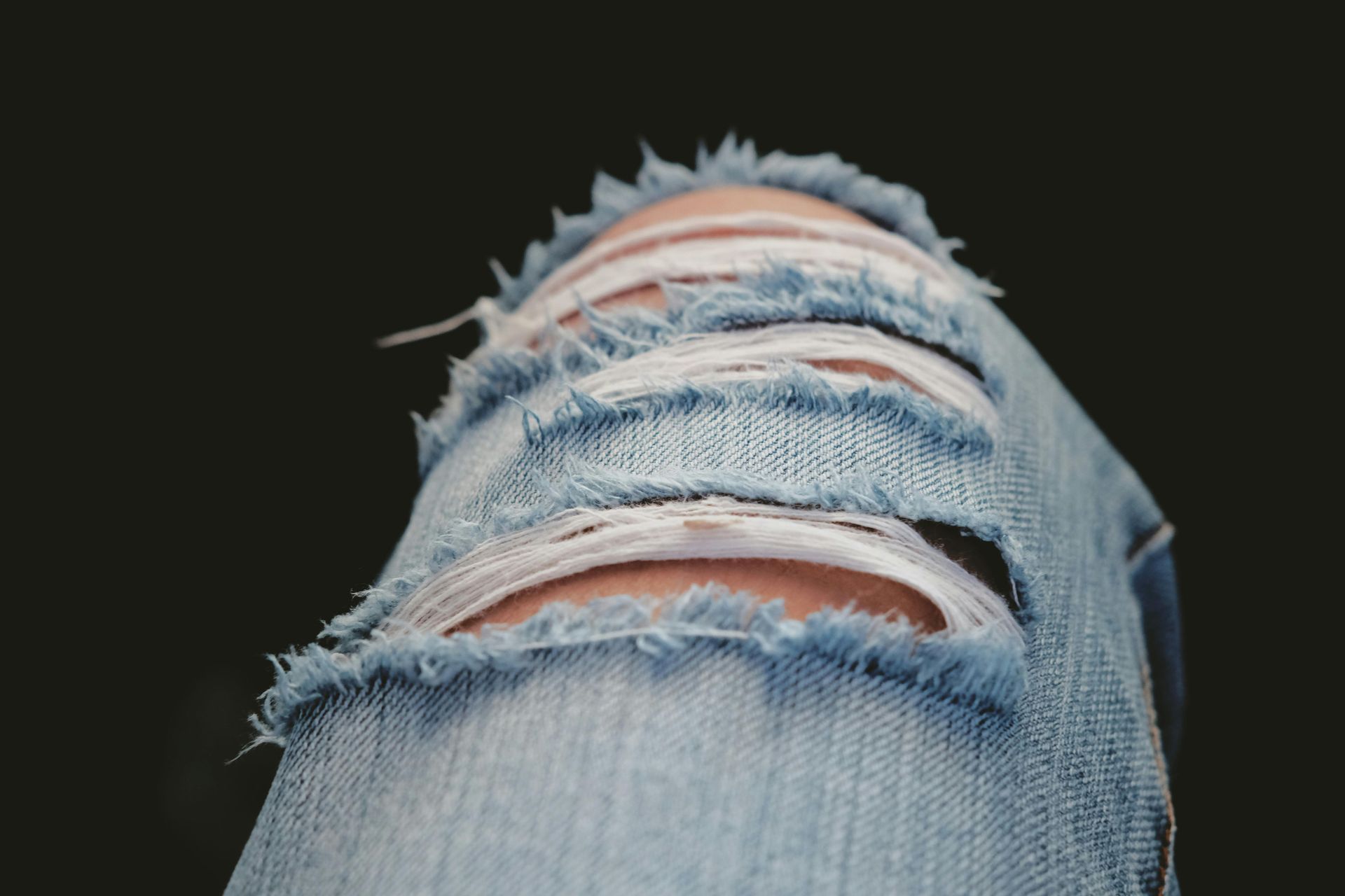 Close-up of ripped blue jeans over a knee, showing skin through the holes, against a black background.