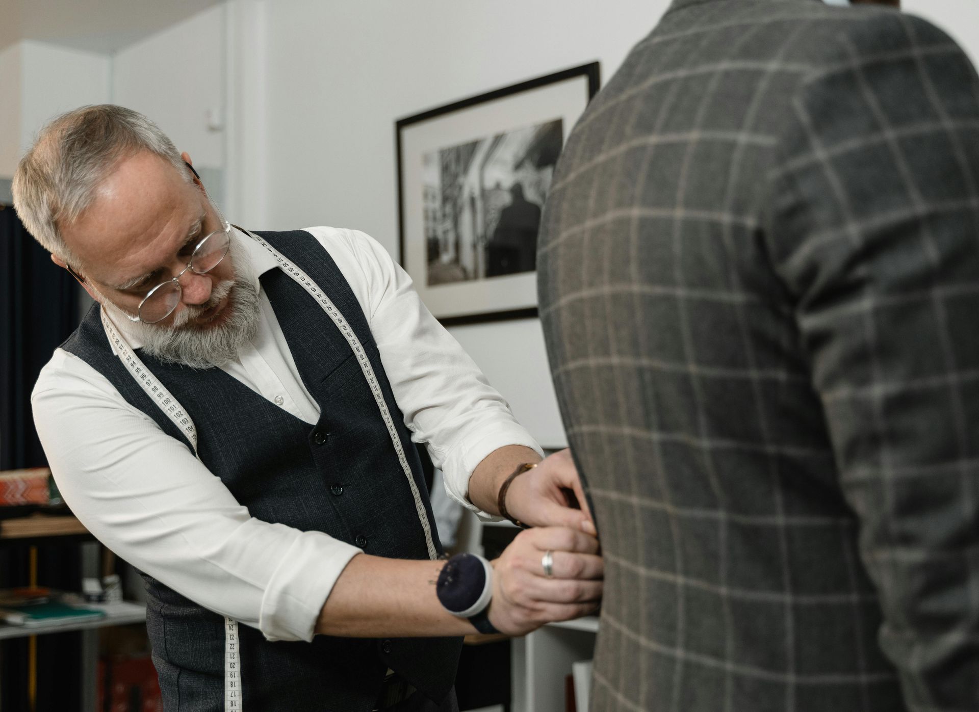 Tailor measures a plaid suit jacket on a person in a workshop.