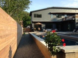 A modern house with a pool, gravel path, and wood fence. Flowers and outdoor seating are visible.