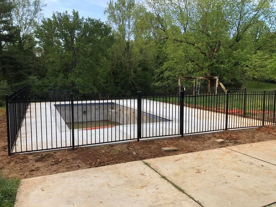 Black fenced-in rectangular pool area surrounded by dirt and concrete, trees in background.