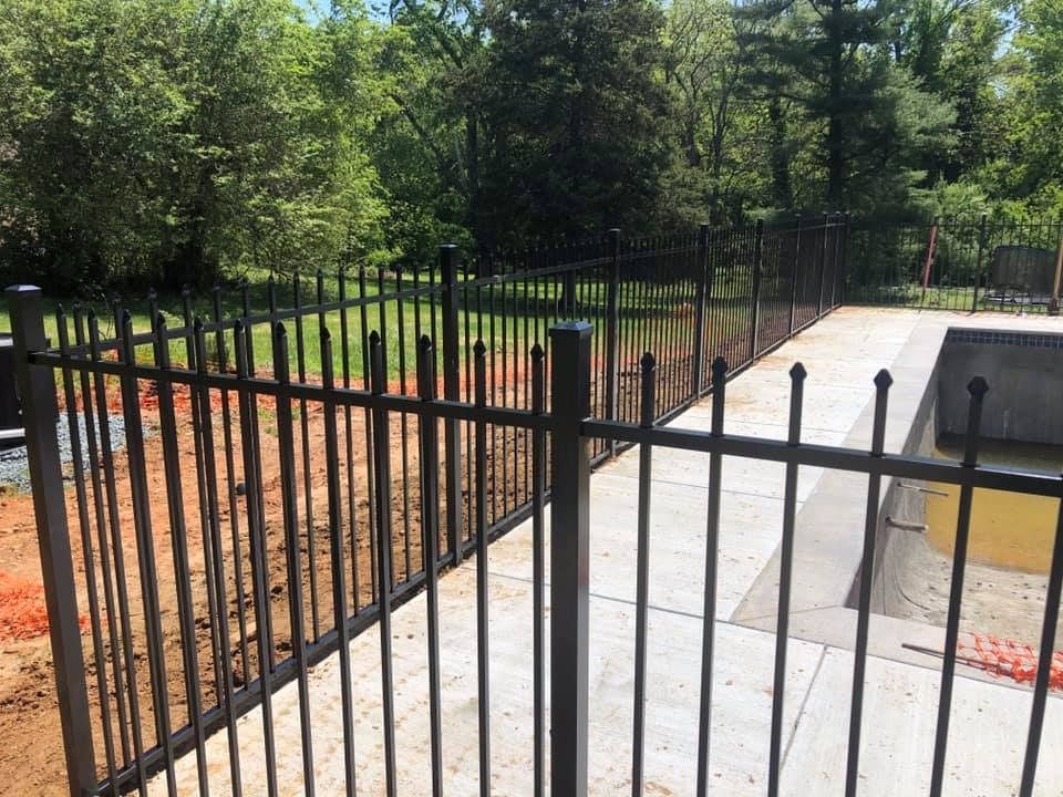 Black metal fence encloses a pool area, viewed outdoors with trees in the background.